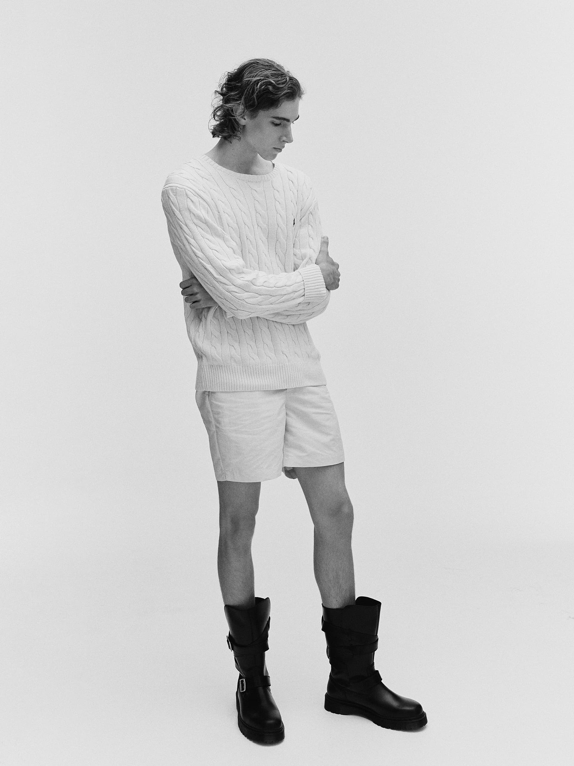 Man wearing a cable-knit sweater, shorts, and boots, in a minimalist black and white style.