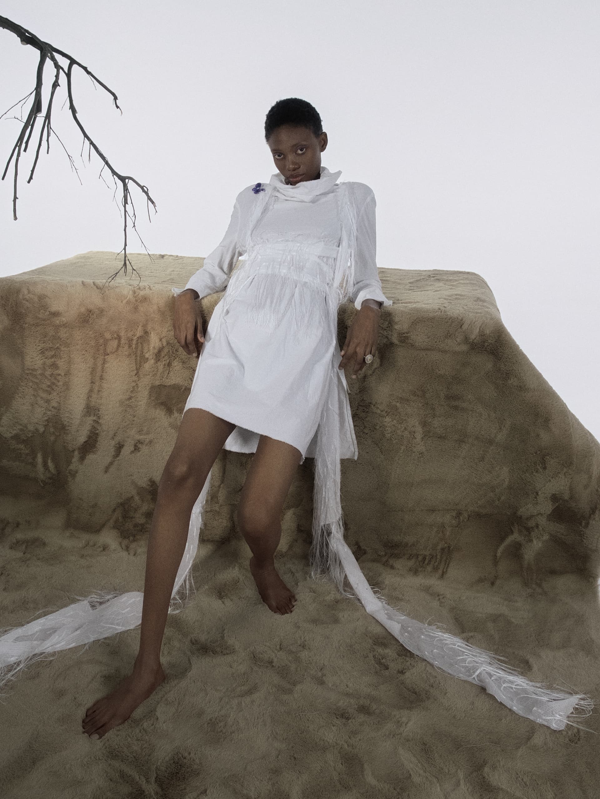 Minimalist fashion shoot: person in white dress on sand, with bare branch backdrop.