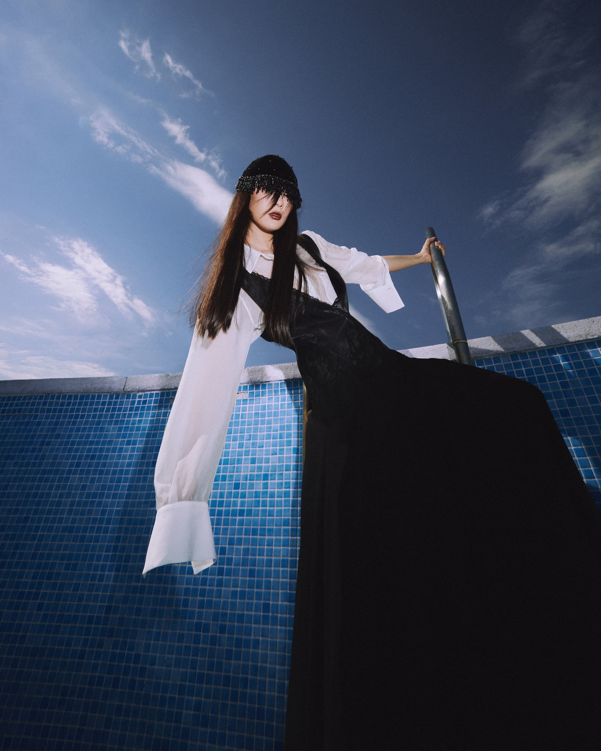 Fashion model posed by a pool in chic black and white outfit under a blue sky.