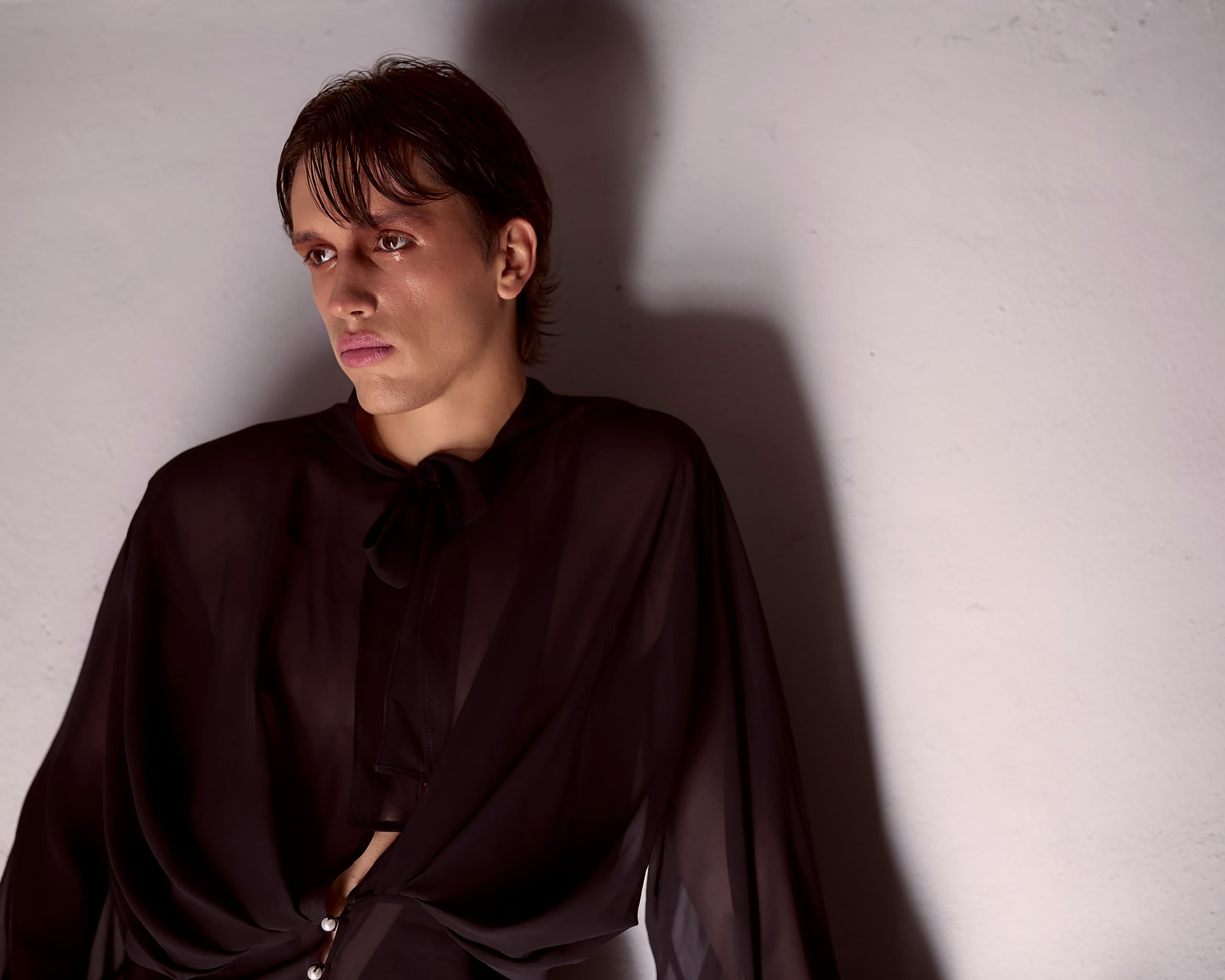 Person in sheer black blouse gazing thoughtfully against a plain white wall.