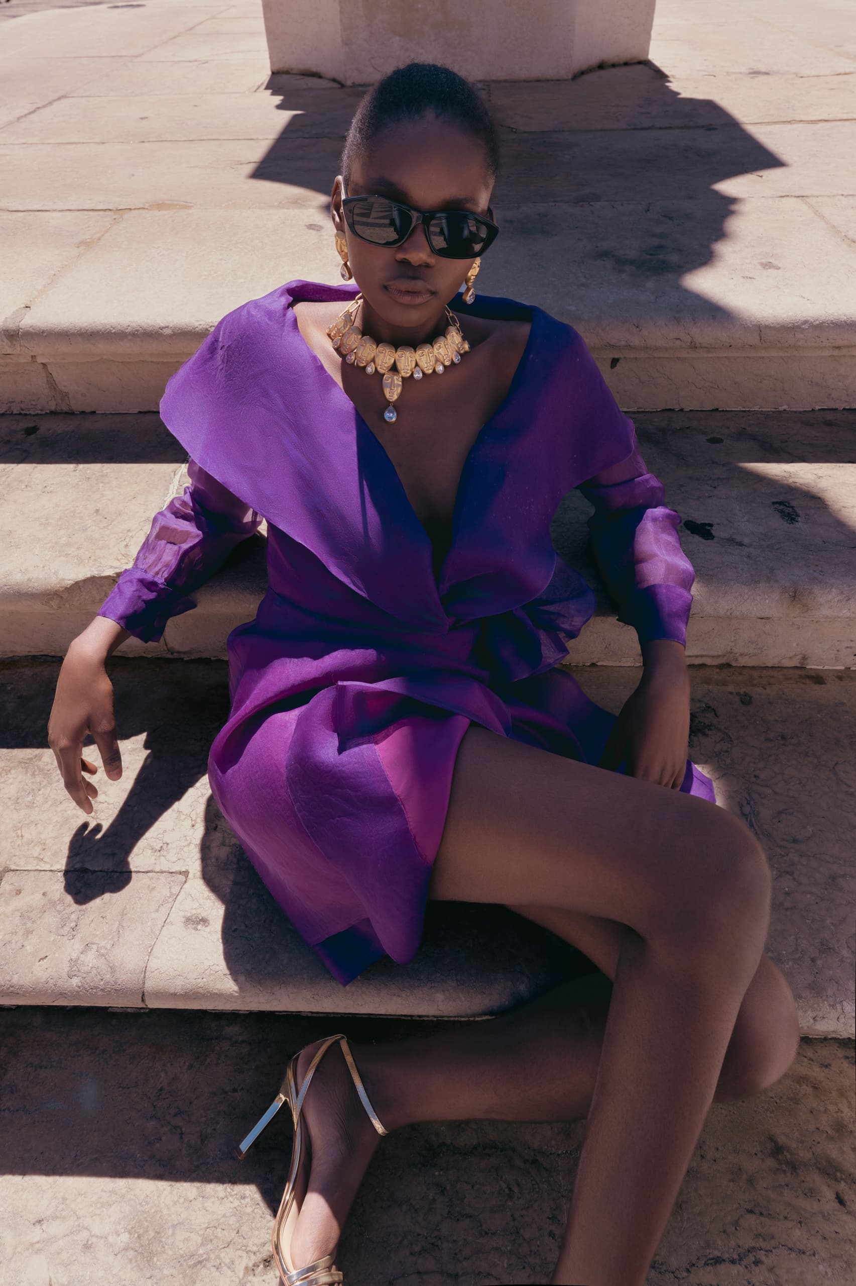 Fashionable woman in purple dress and gold accessories, sitting on steps in sunlight.