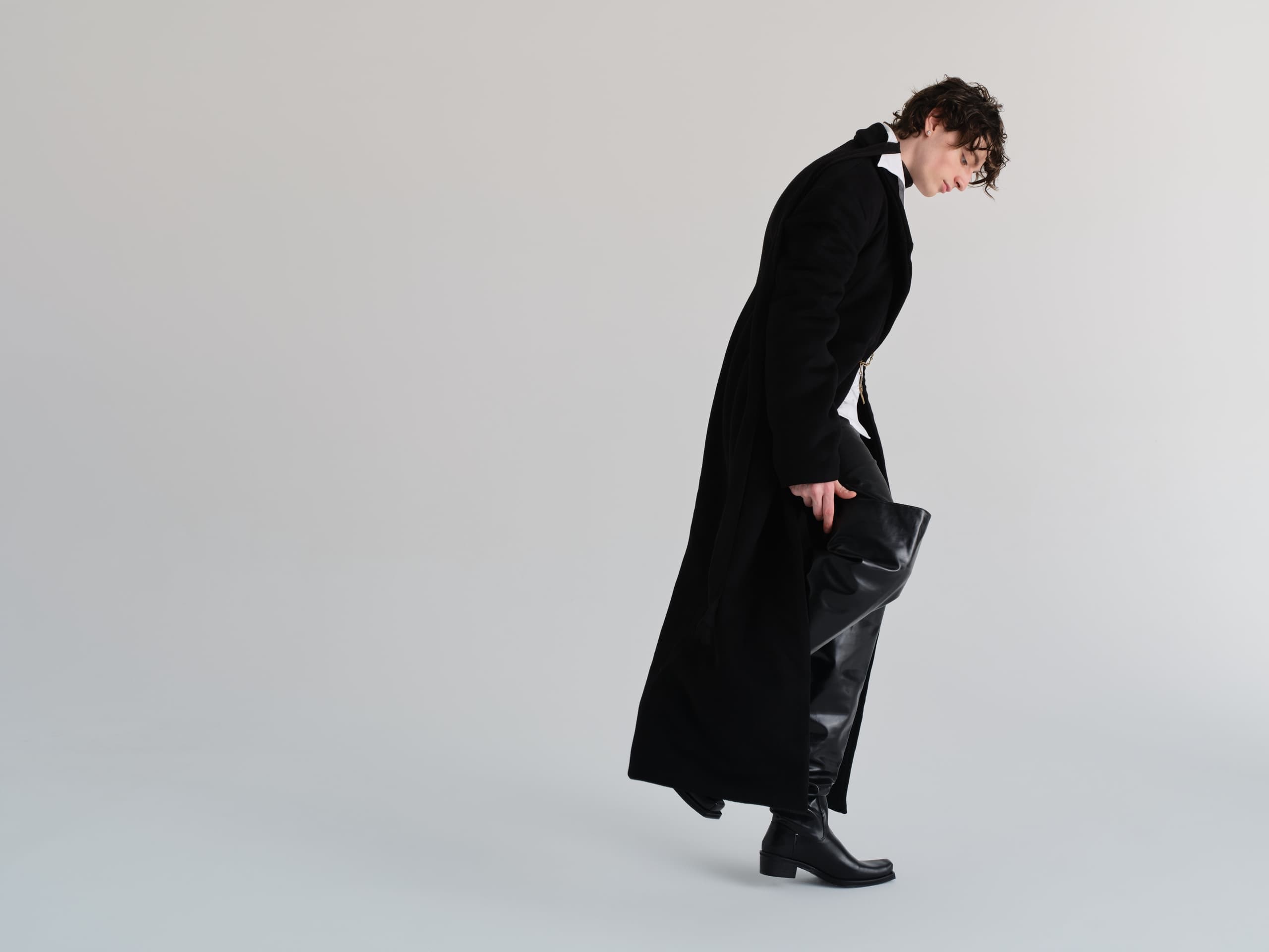 Person in black coat and boots posing against a minimalist background.
