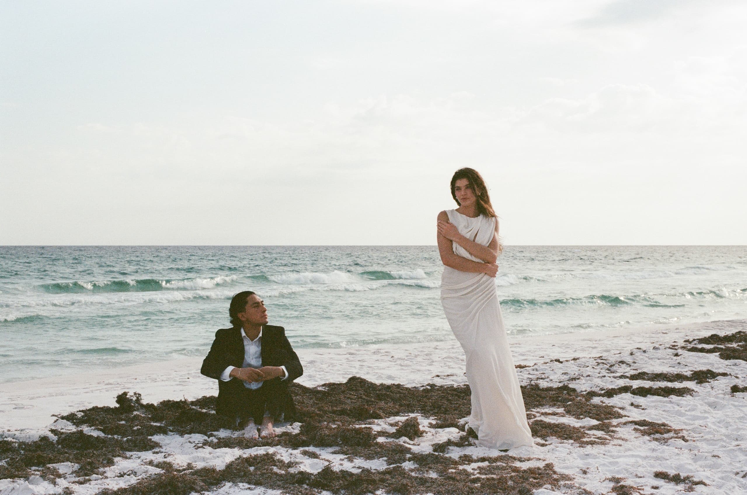 Couple on a beach at sunset, one seated, one standing in elegant attire.