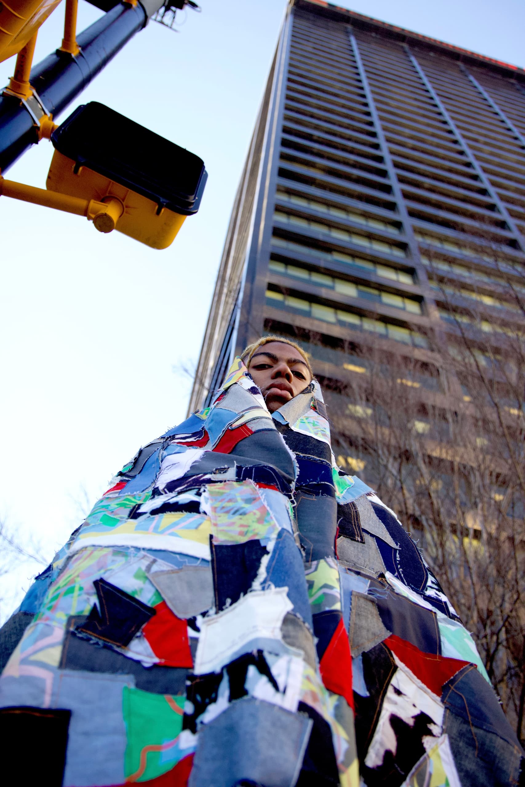 Person draped in colorful patchwork near modern skyscraper and traffic light.