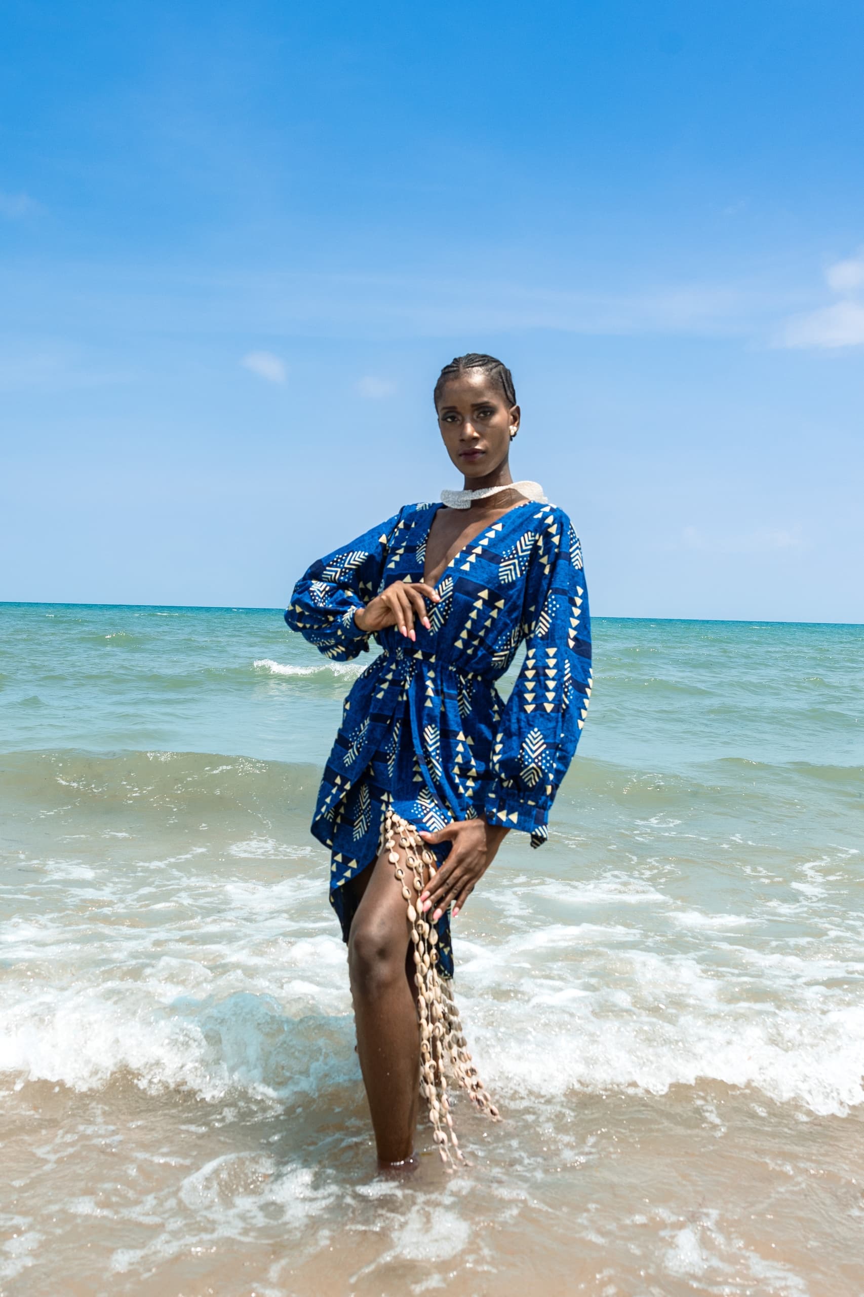 Model in blue patterned dress poses by the ocean under clear skies.