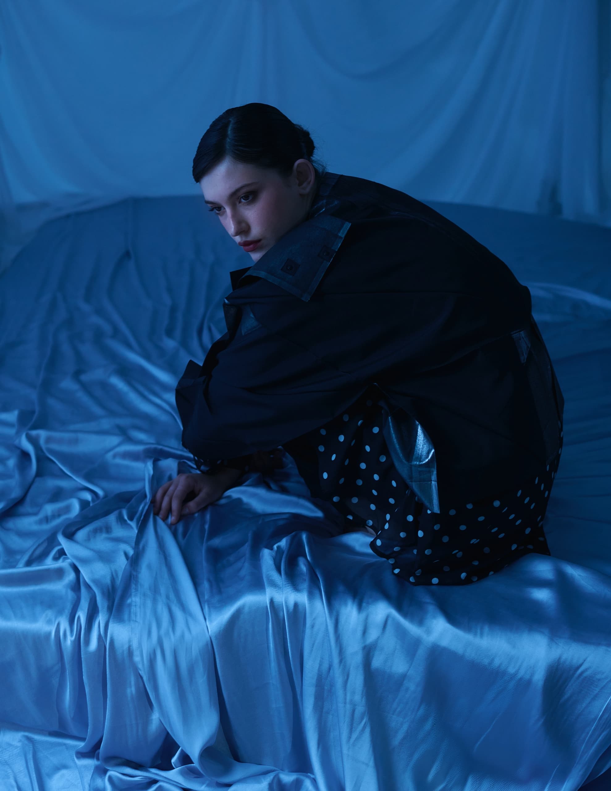 Person in dark coat on blue silk bed, moody lighting.