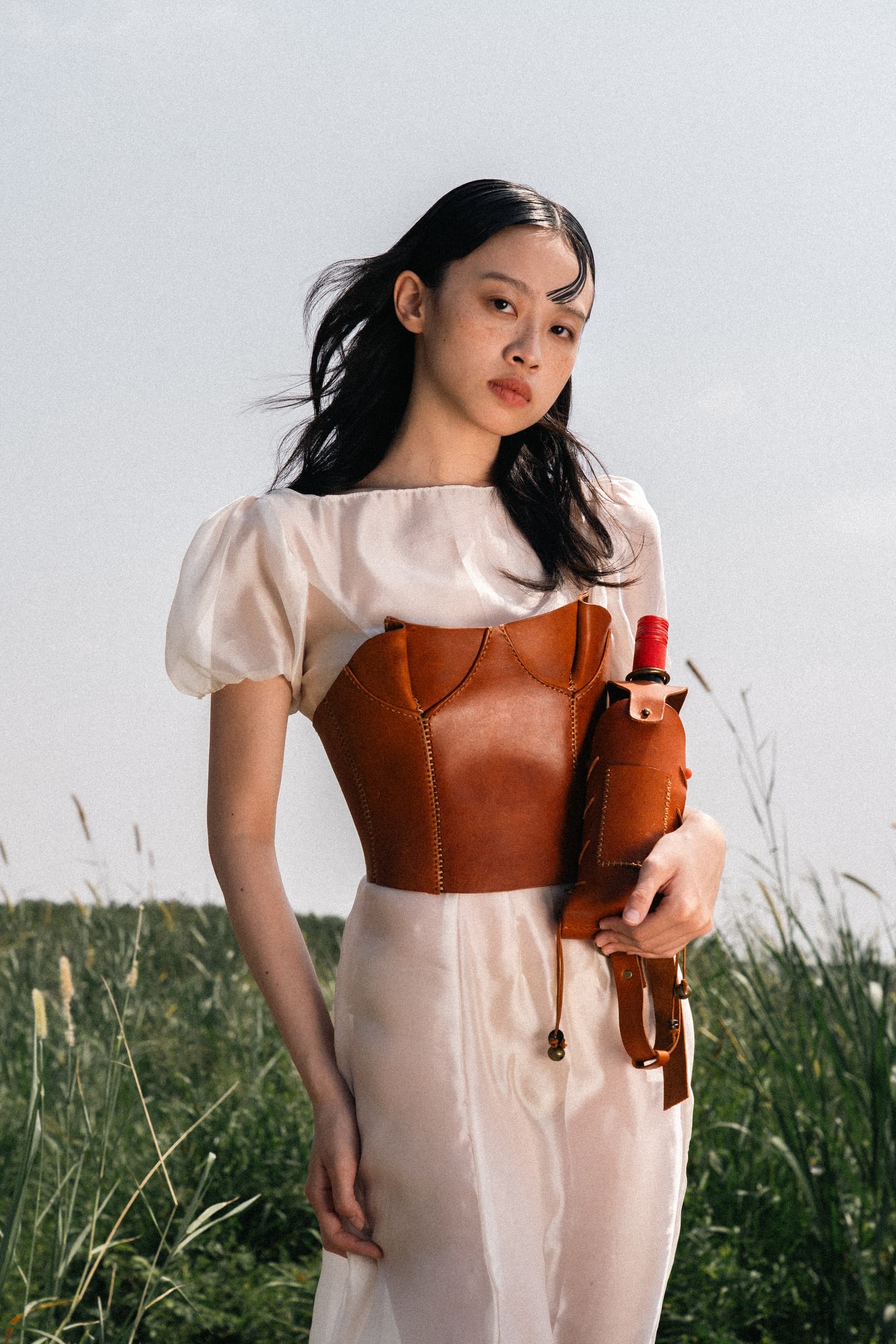 Woman in white dress and leather corset outdoors, holding a wine bottle.