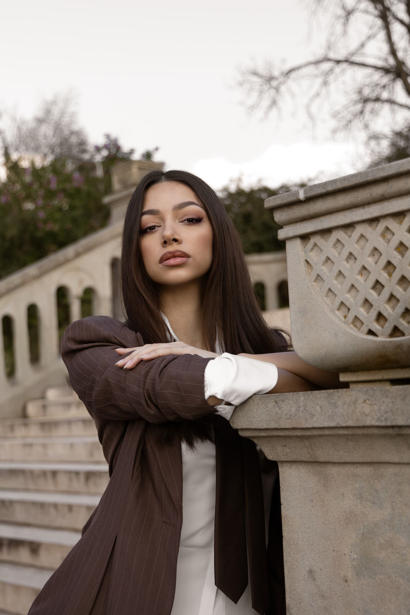 Stylish woman in a brown suit poses elegantly near stone staircase outdoors.
