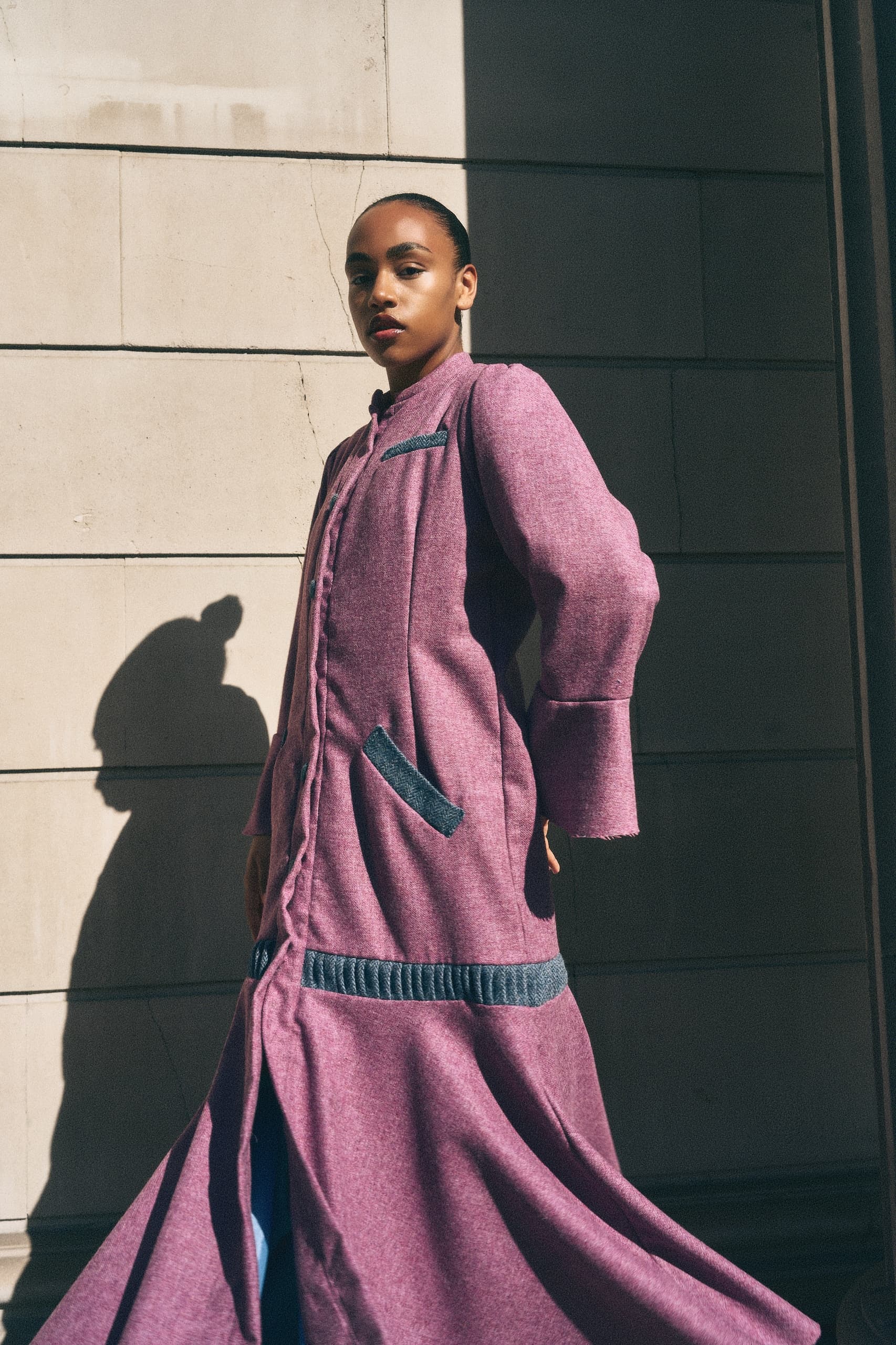 Model in stylish pink coat poses against sunlit urban wall.