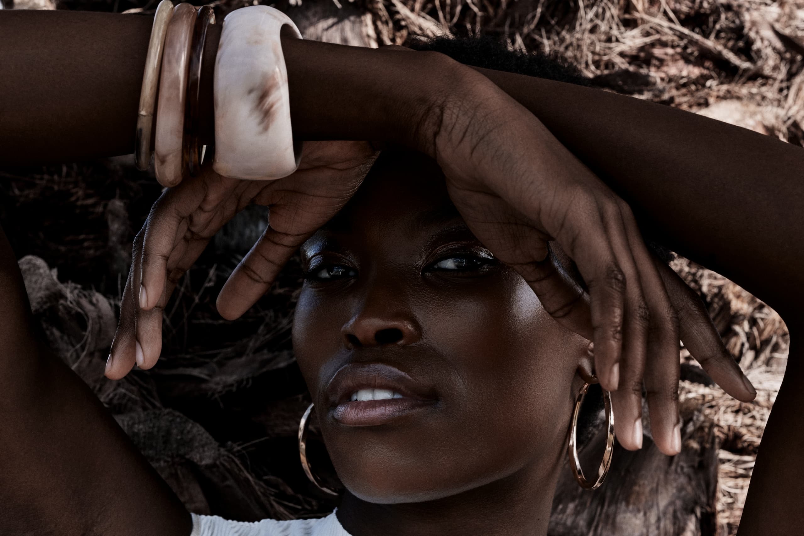 Woman poses with bracelets under dappled sunlight, wearing large hoop earrings.