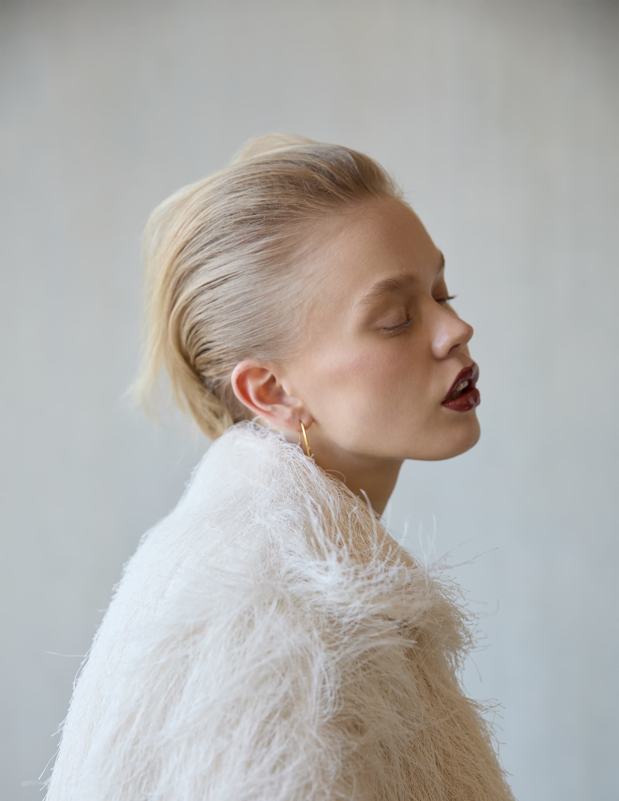 Woman in white feather coat with slicked-back hair and gold hoop earring.
