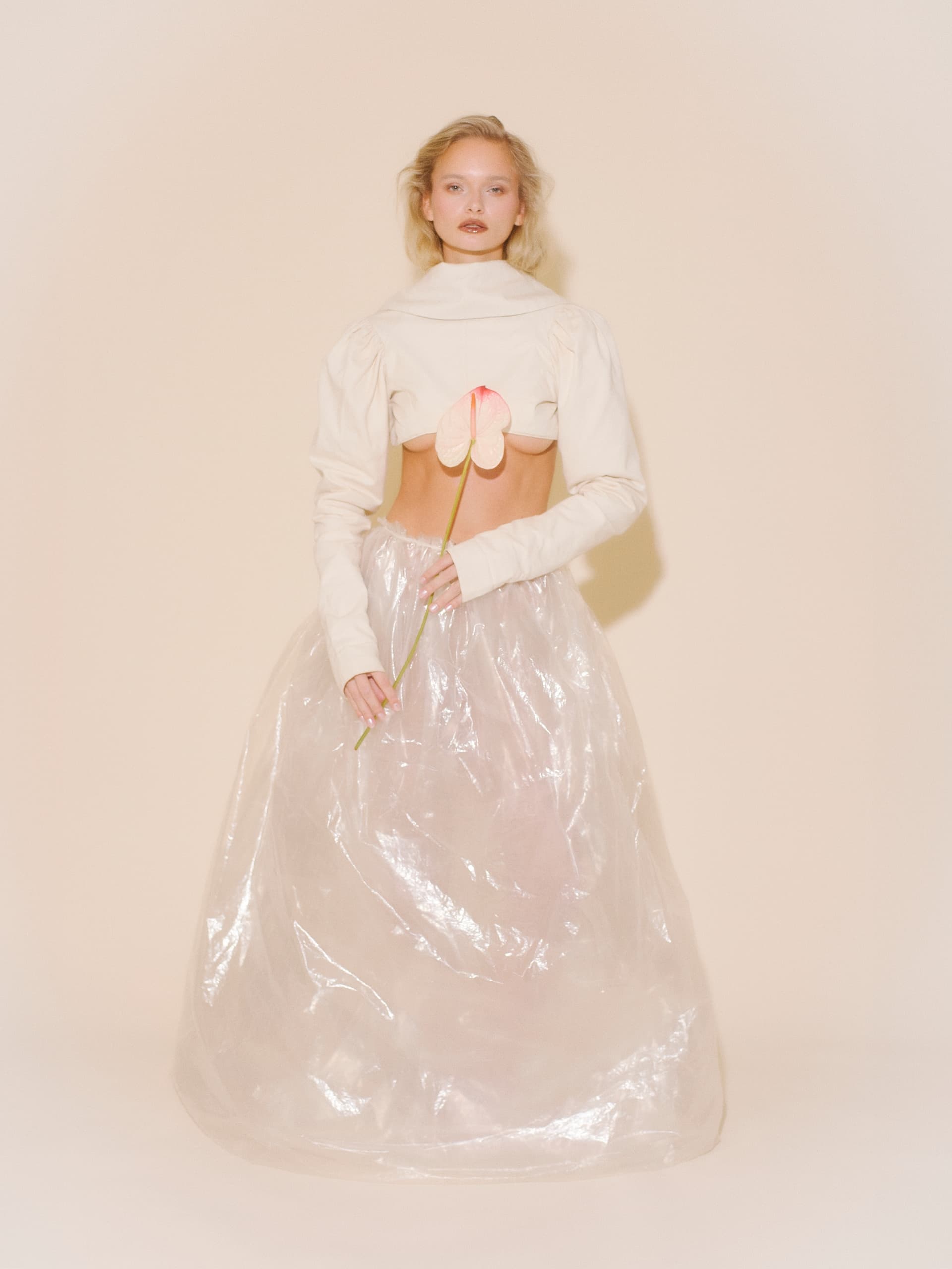 Elegant woman in a sheer skirt holding a pink flower in a minimalist setting.