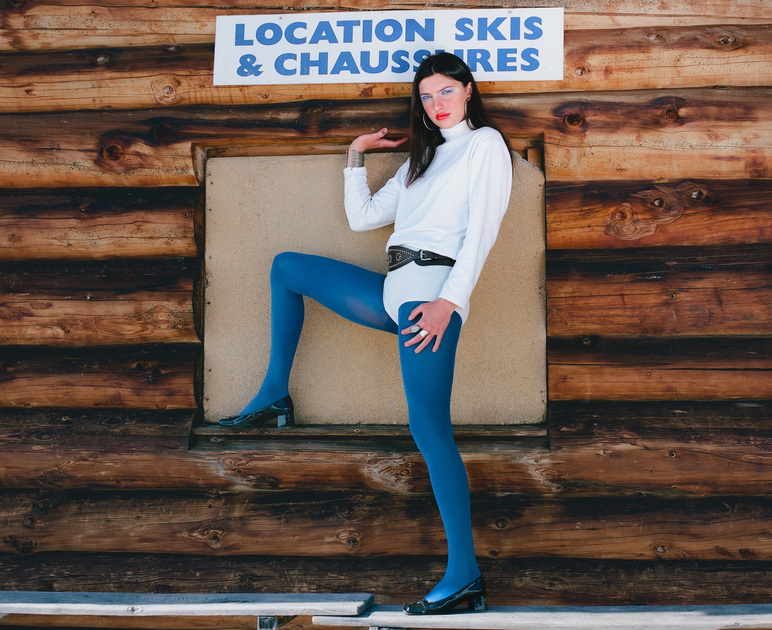Woman in stylish ski lodge outfit poses by wooden building with ski rental sign.
