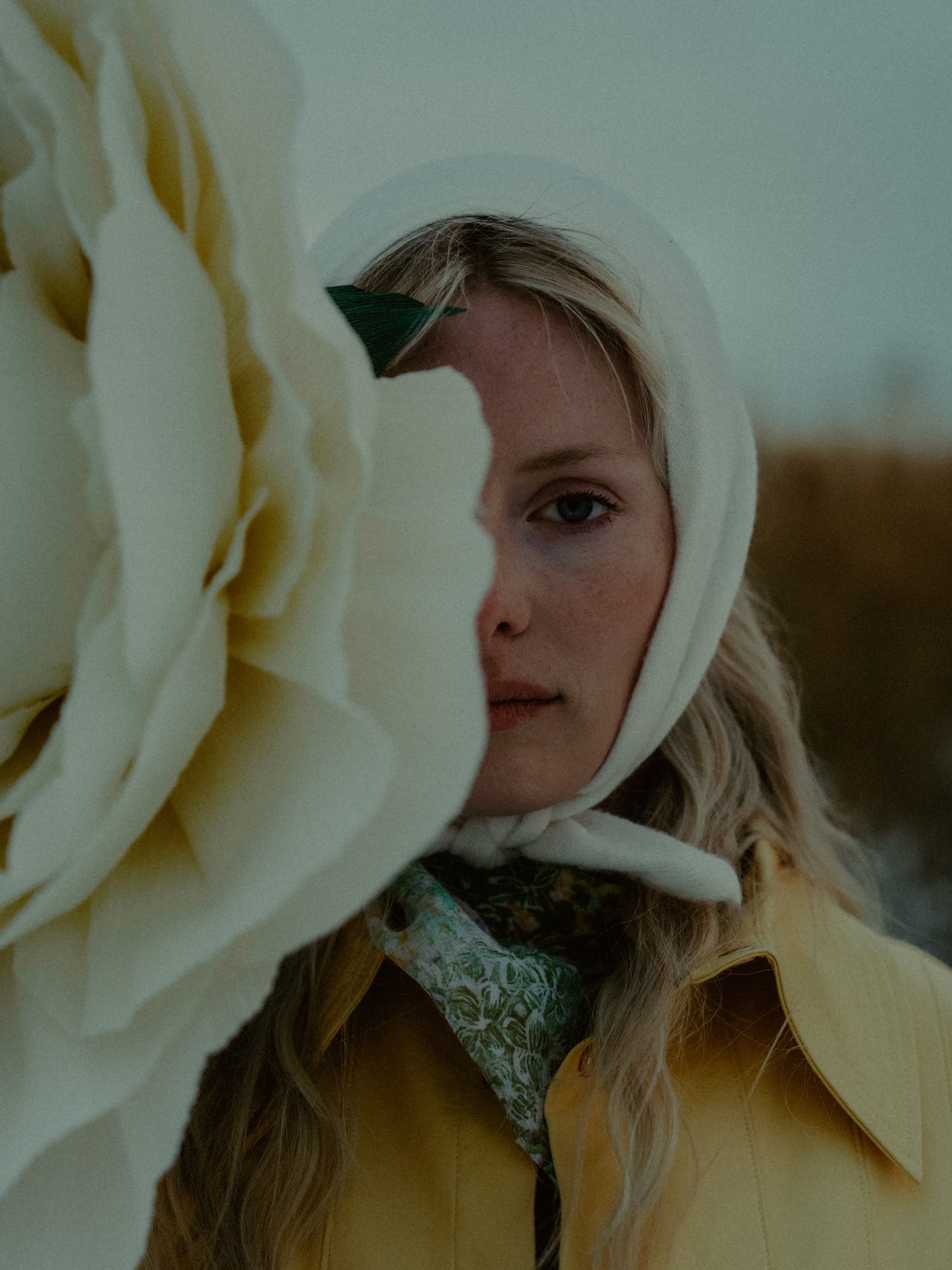 Woman in a white headscarf with giant white flower, outdoors in soft light.