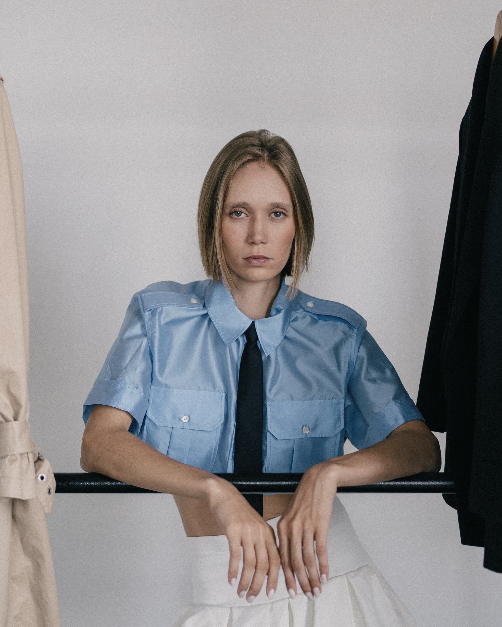 Woman in blue shirt with tie, leaning on clothes rack, stylish fashion portrait.
