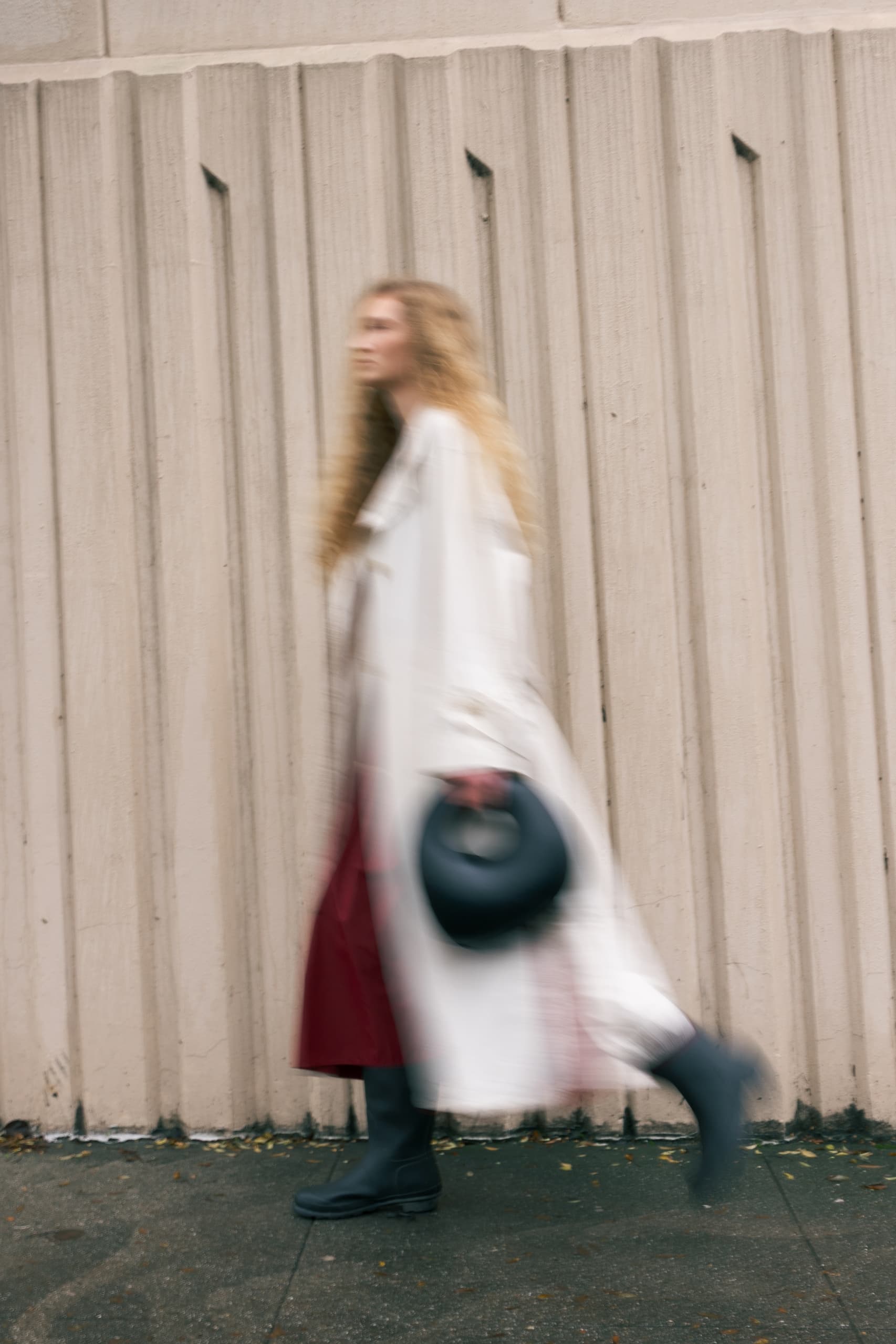 Blurred person walking by a textured wall, holding a hat, wearing a white coat.