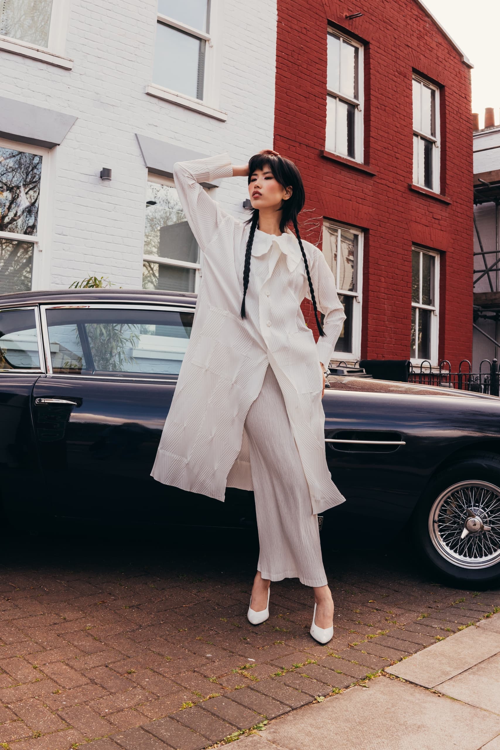 Fashionable woman posing in white outfit by classic car outside urban building.