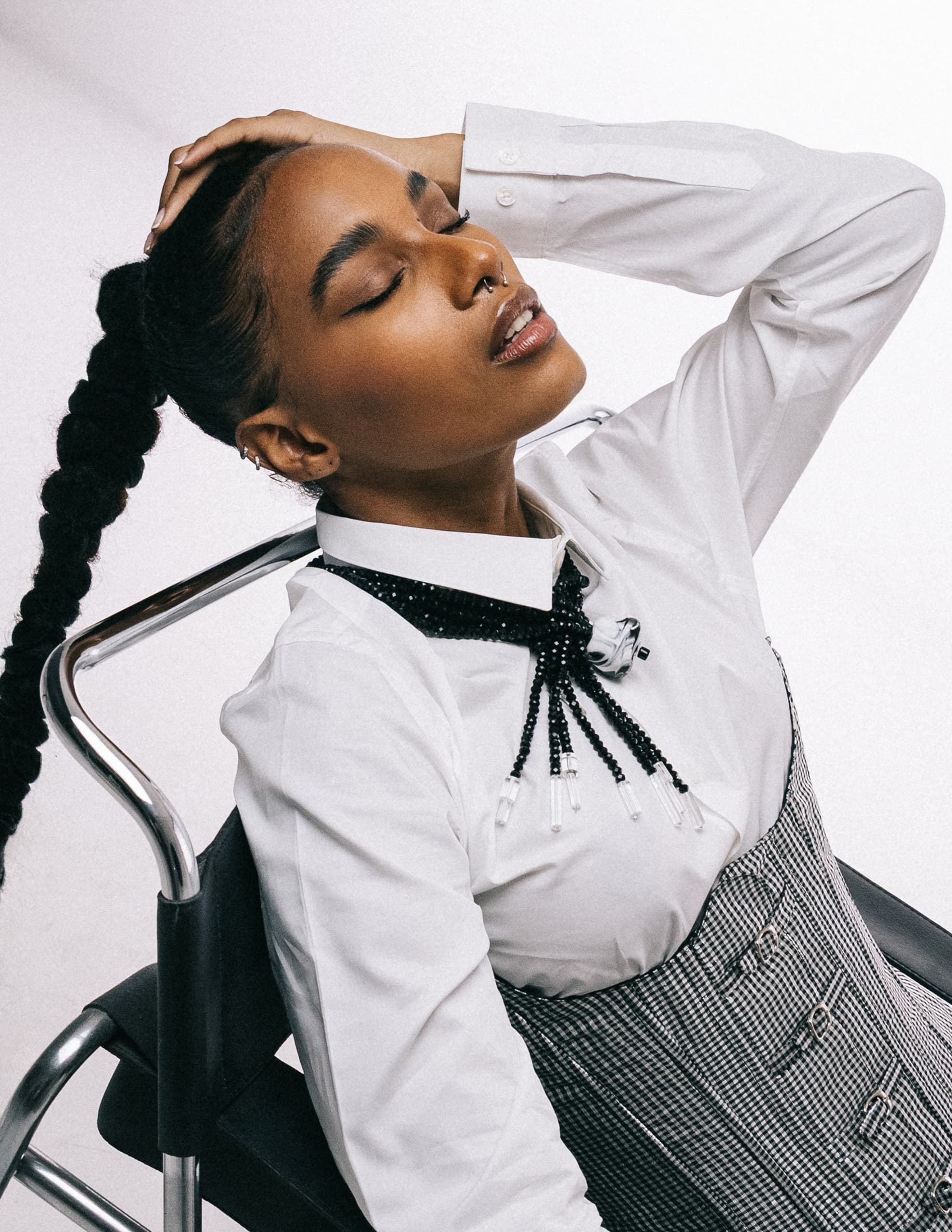 Fashionable person with braid, corset, and white shirt posing on chair.