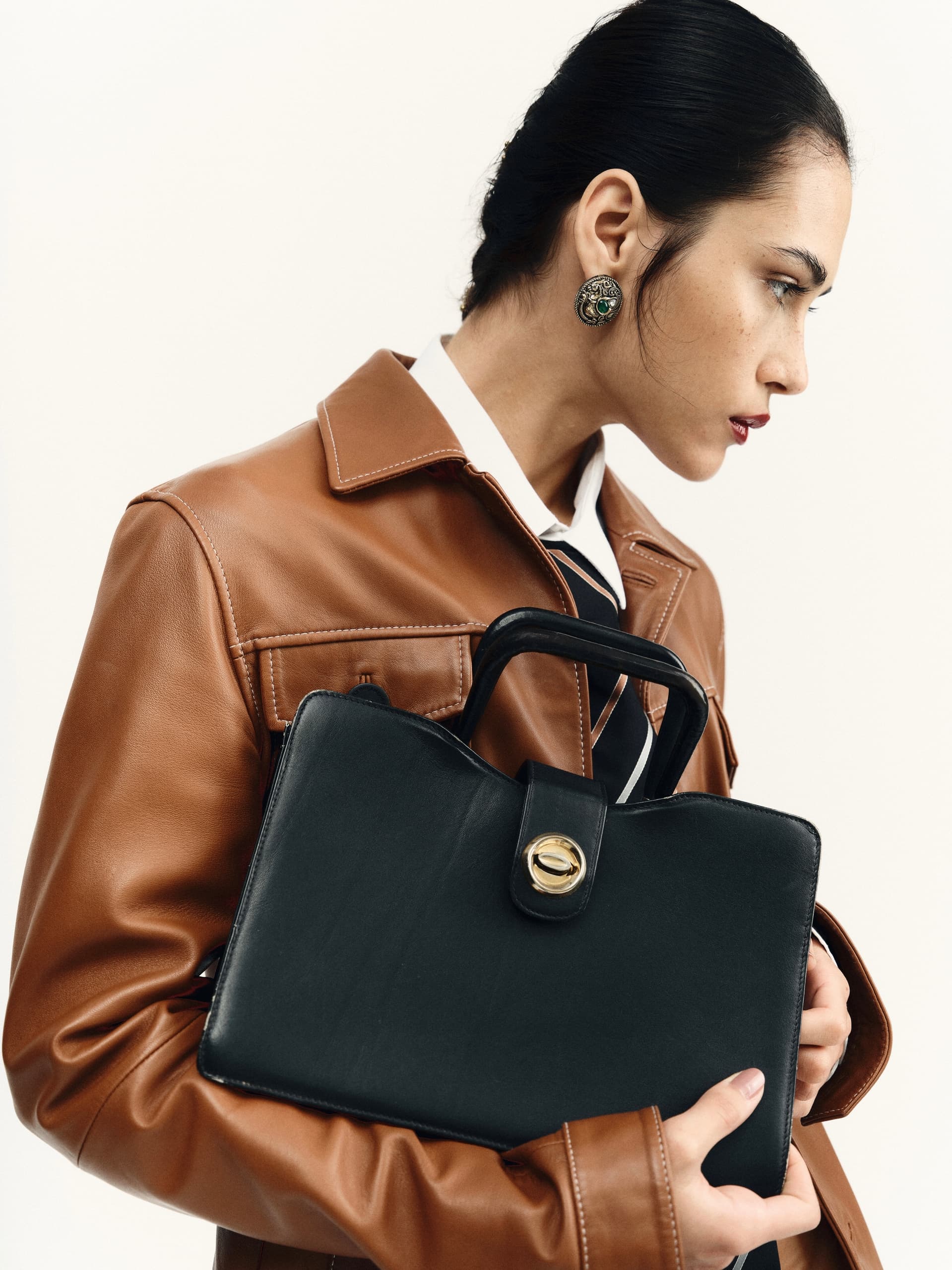 Woman in brown jacket holding a black handbag, showcasing modern fashion style.