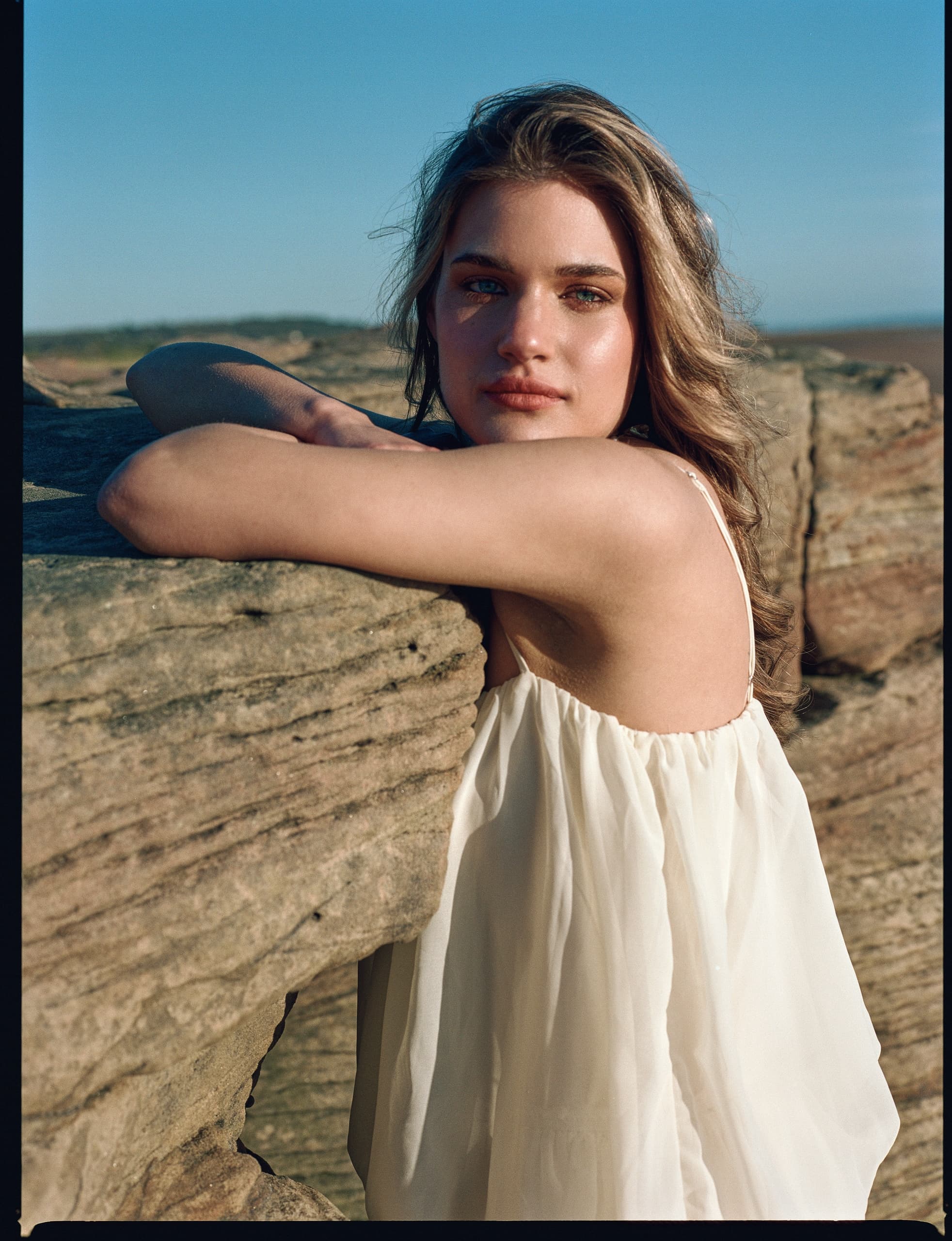 Woman in white dress leaning on rocks under clear blue sky.