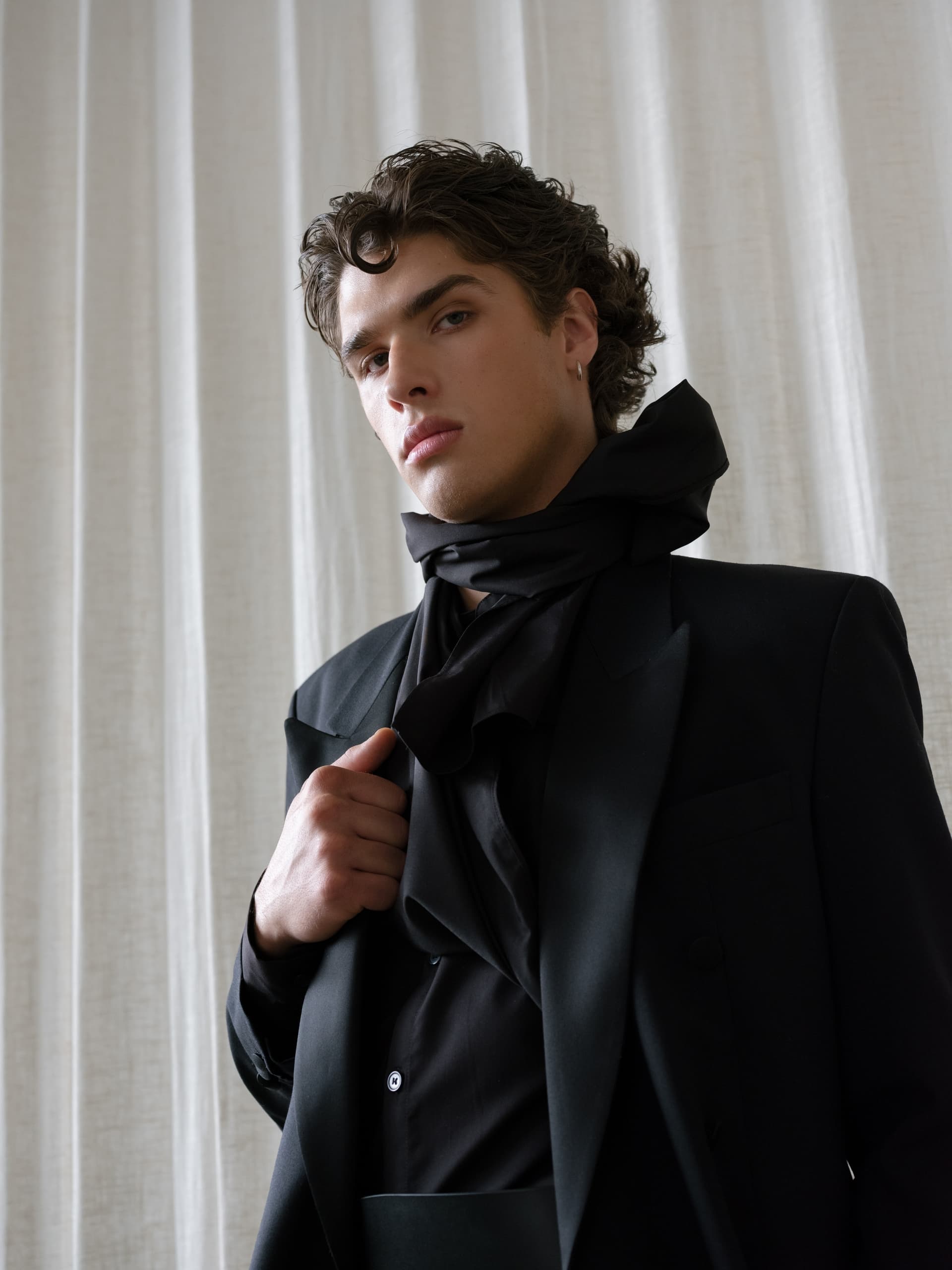 Fashionable man in black suit and scarf against a light curtain backdrop.