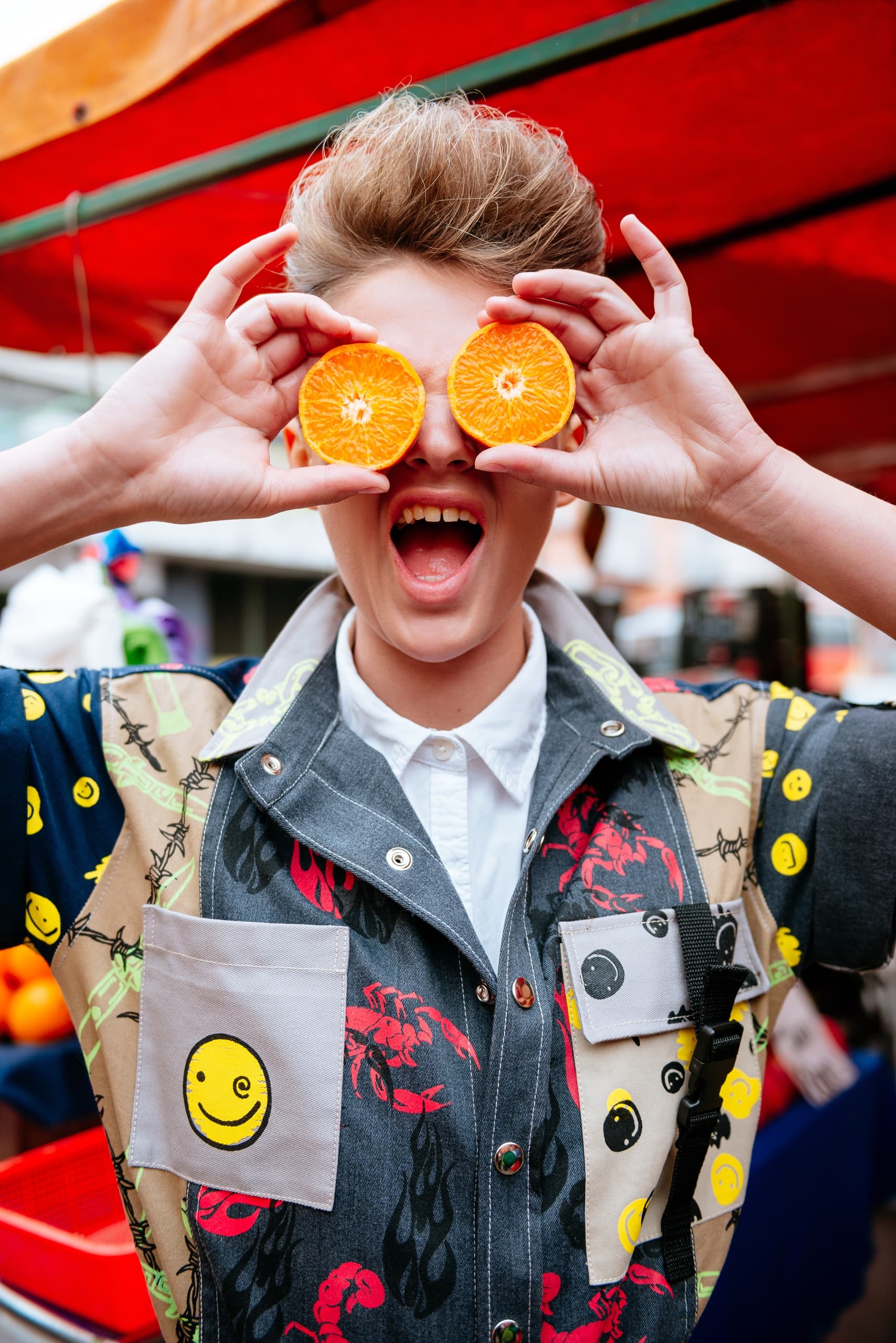 Person playfully holding orange slices over eyes, wearing a colorful jacket.