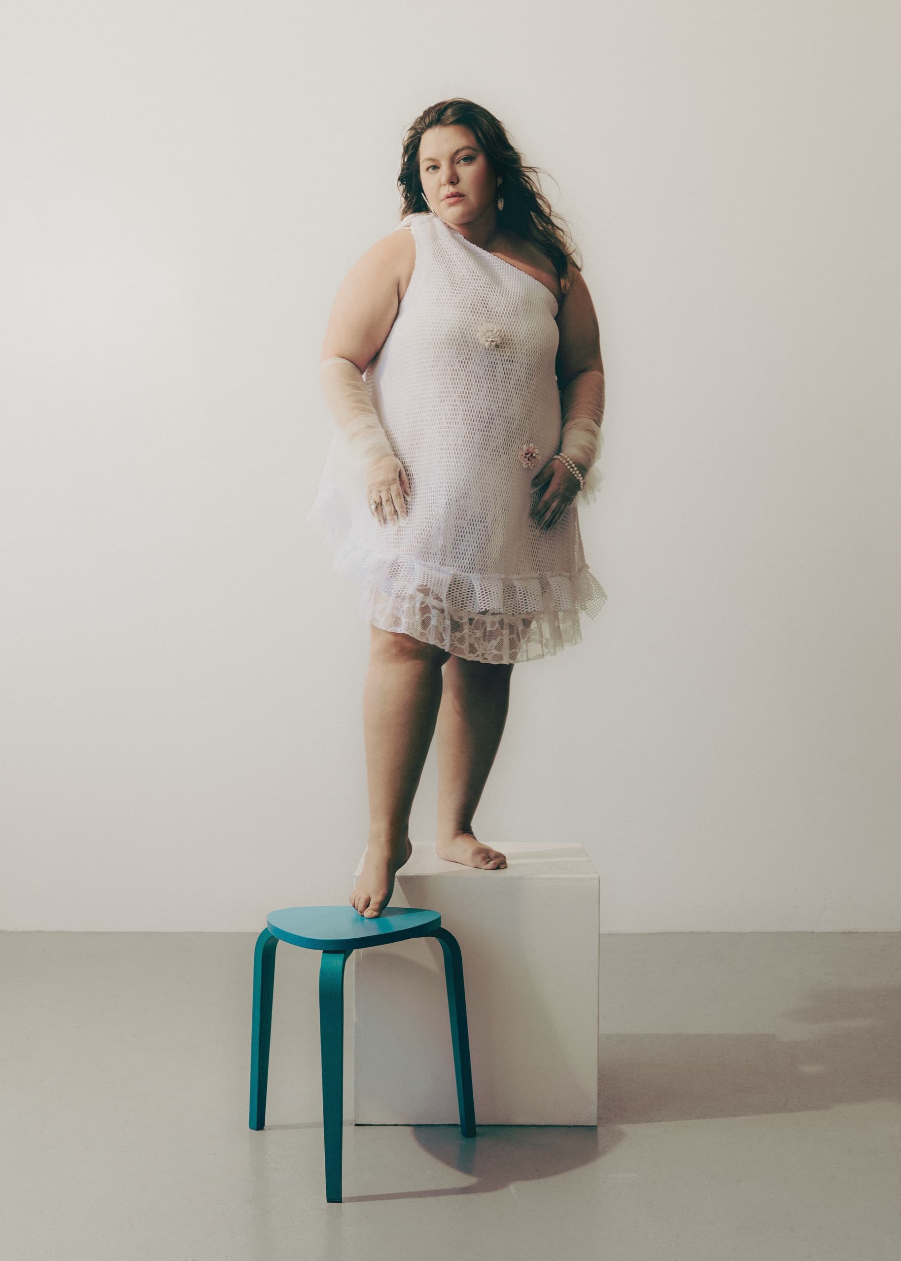 Woman in stylish white dress standing on stool, minimalist background.