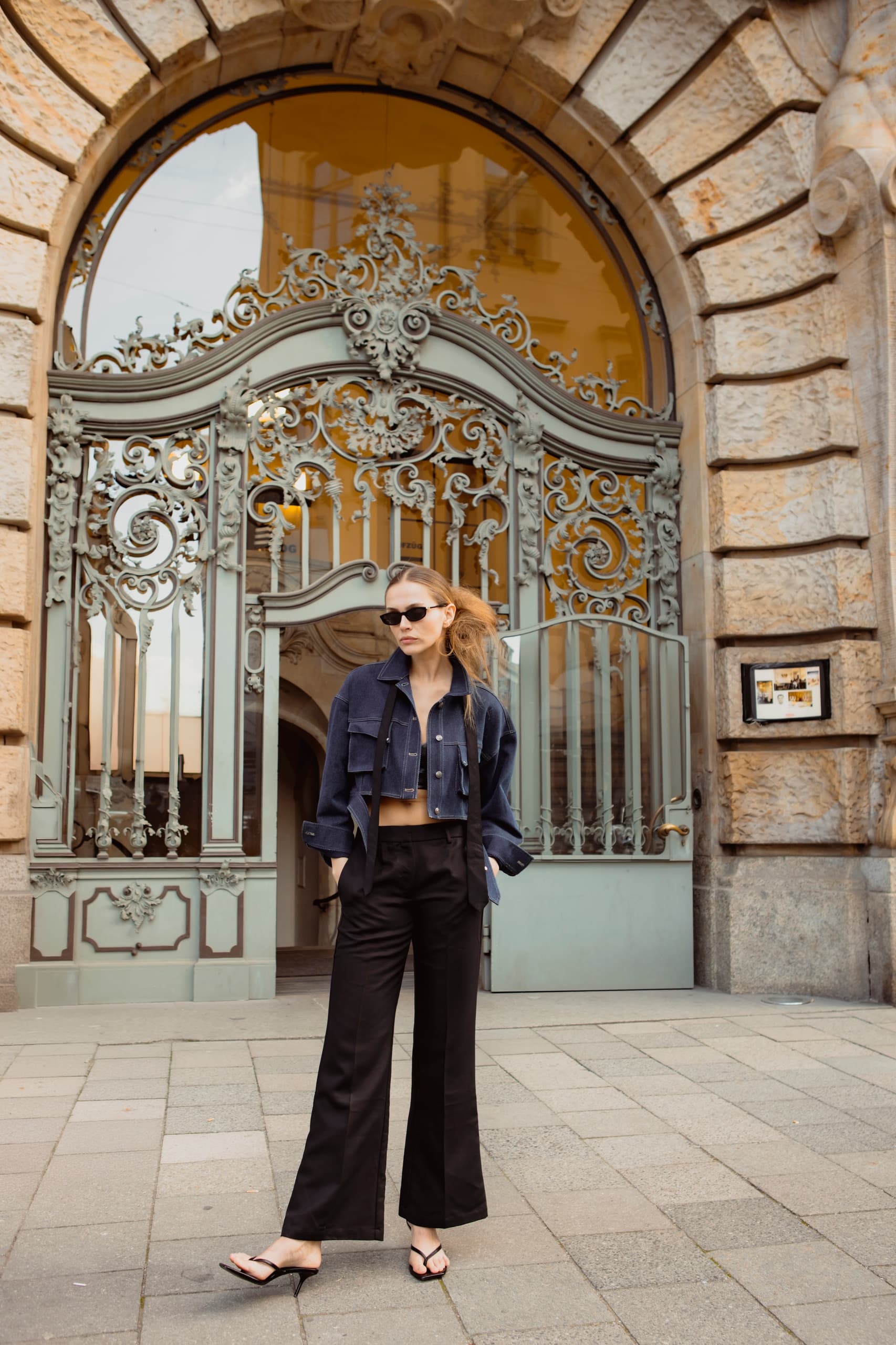 Fashionable woman in front of ornate gate, wearing chic outfit and sunglasses.