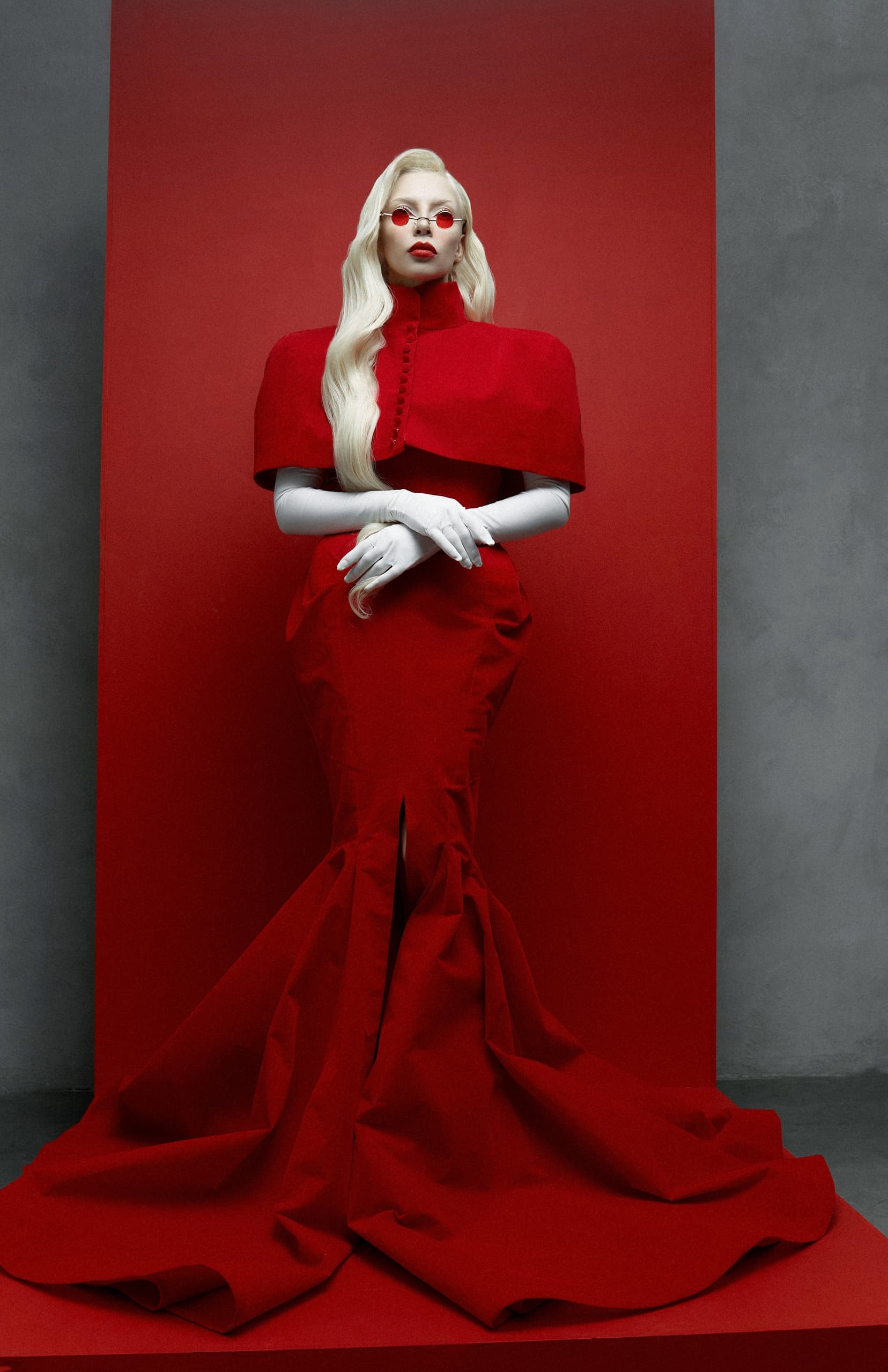 Person in elegant red gown with white gloves against a red and gray backdrop.