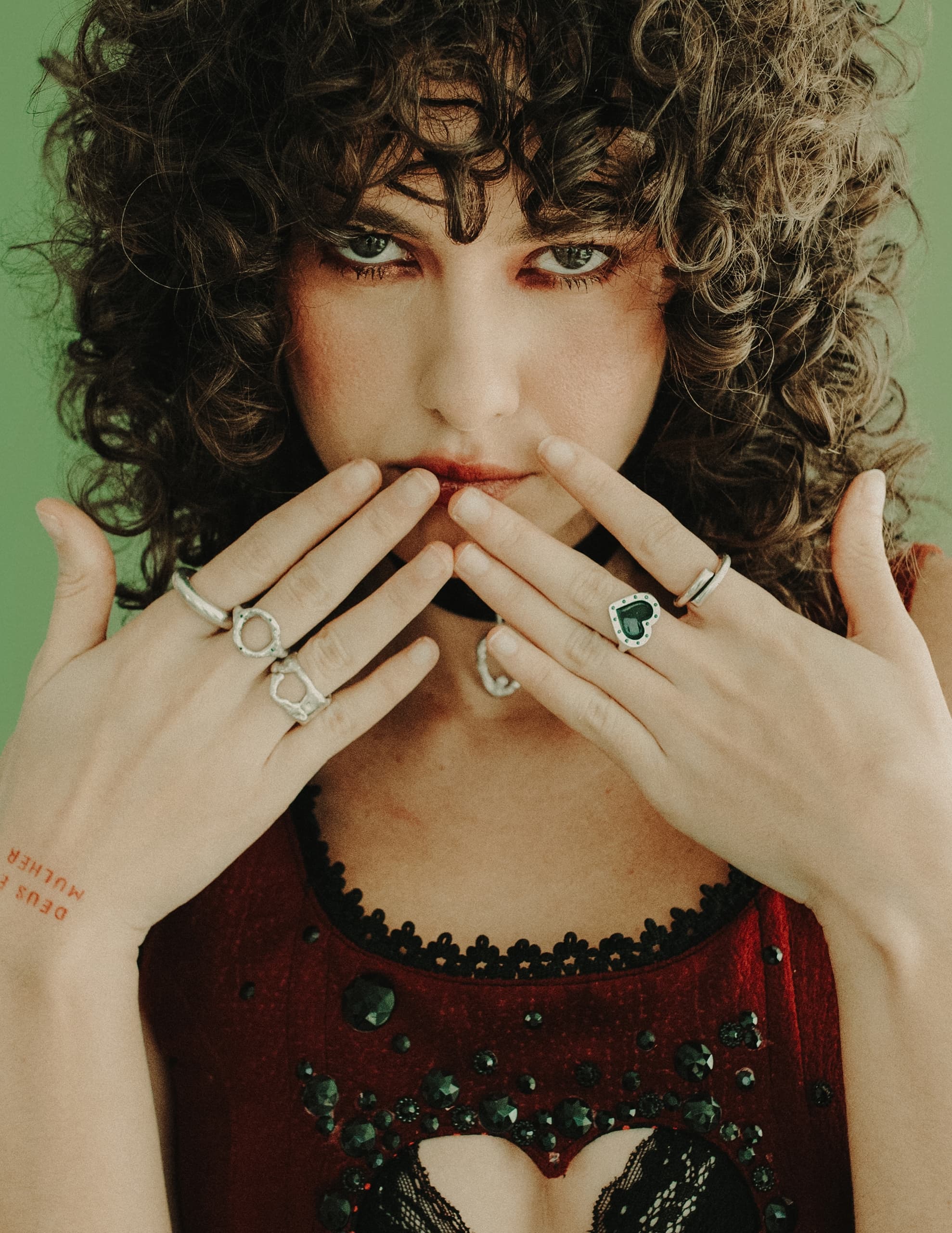 Woman with curly hair and rings, wearing red sequined top, green background.