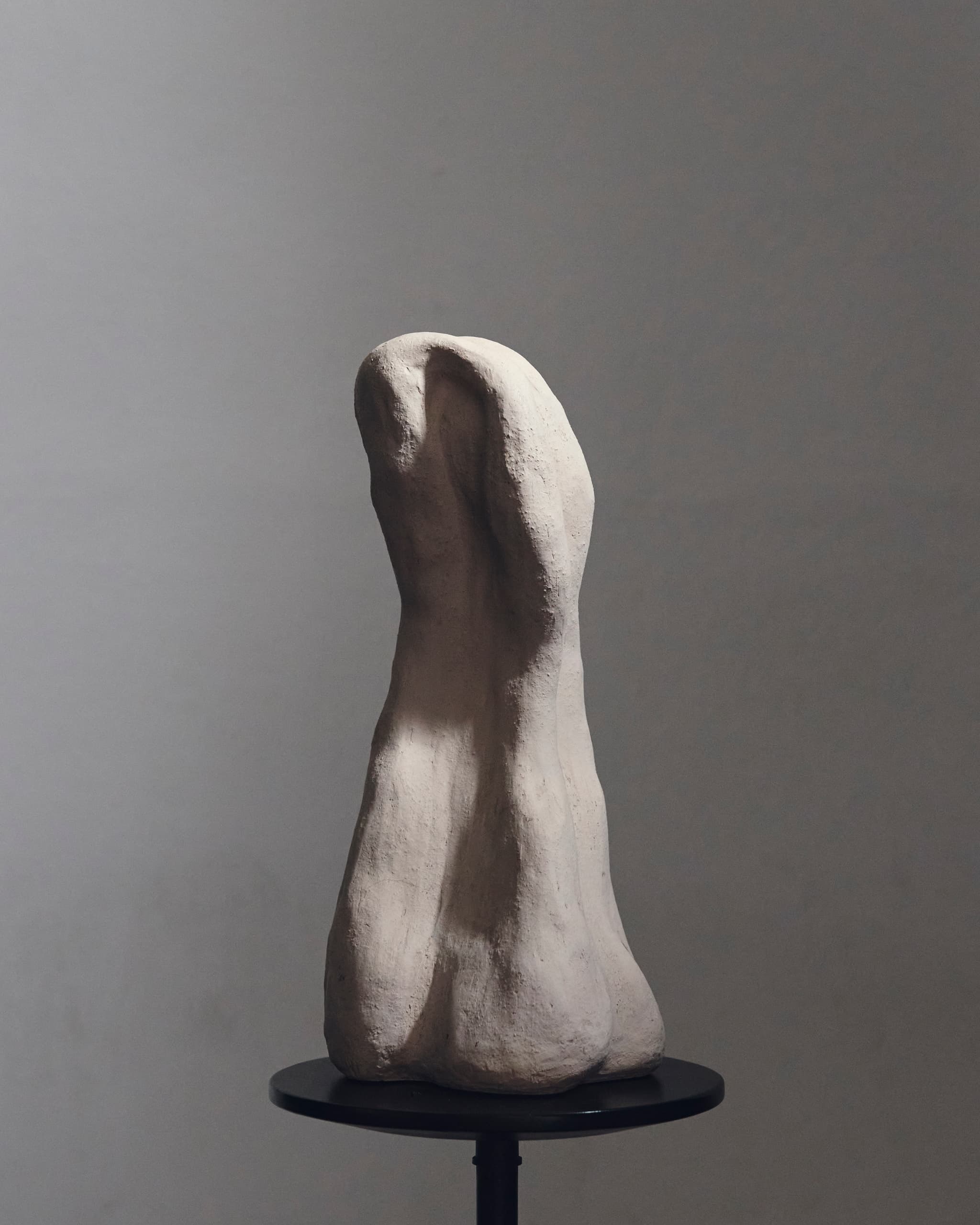 Abstract clay sculpture on dark round pedestal under soft lighting.