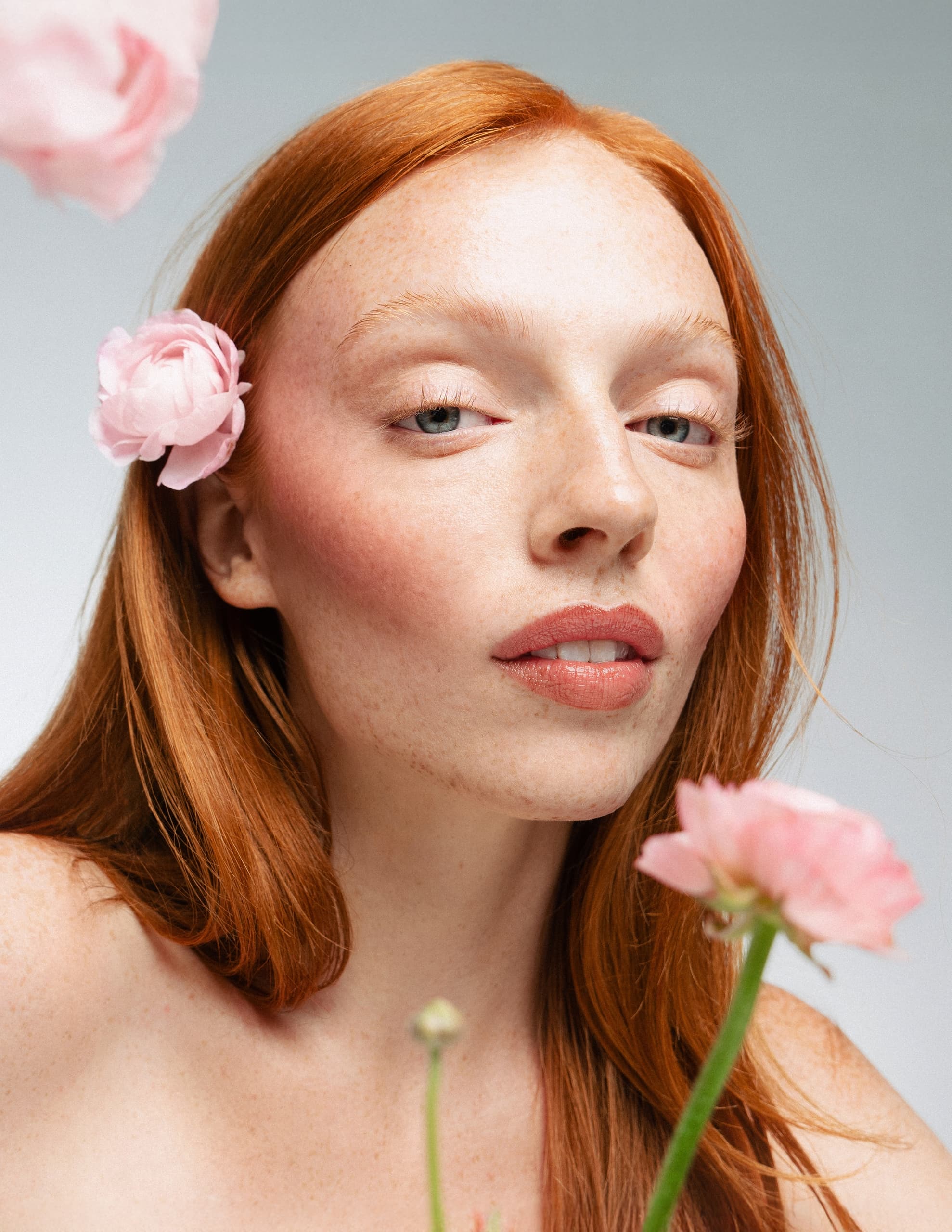 Red-haired woman with pink flowers, serene expression against a neutral background.