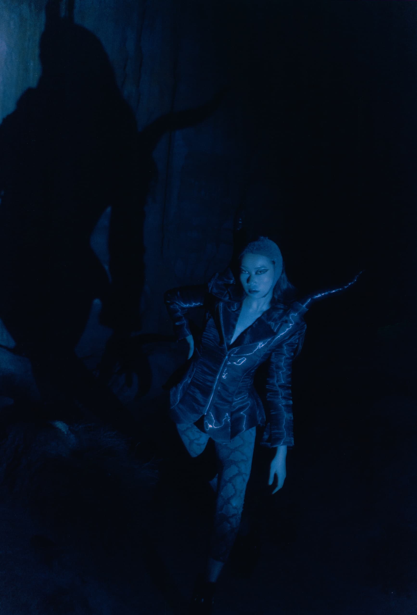 Mysterious figure in shadowy blue light, wearing a jacket with horns and patterned tights.