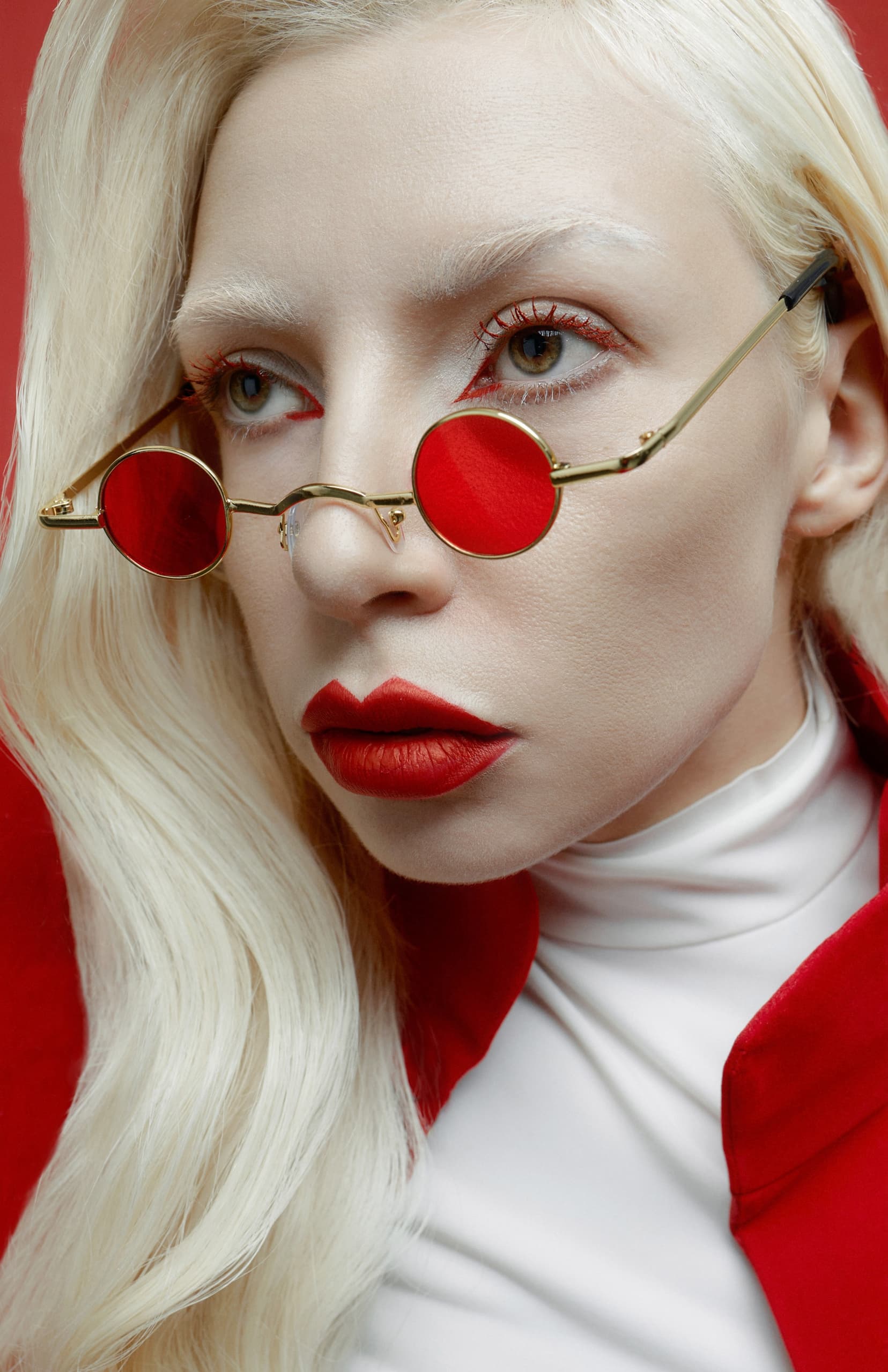 Woman with red glasses and lips, wears a red coat and white shirt.
