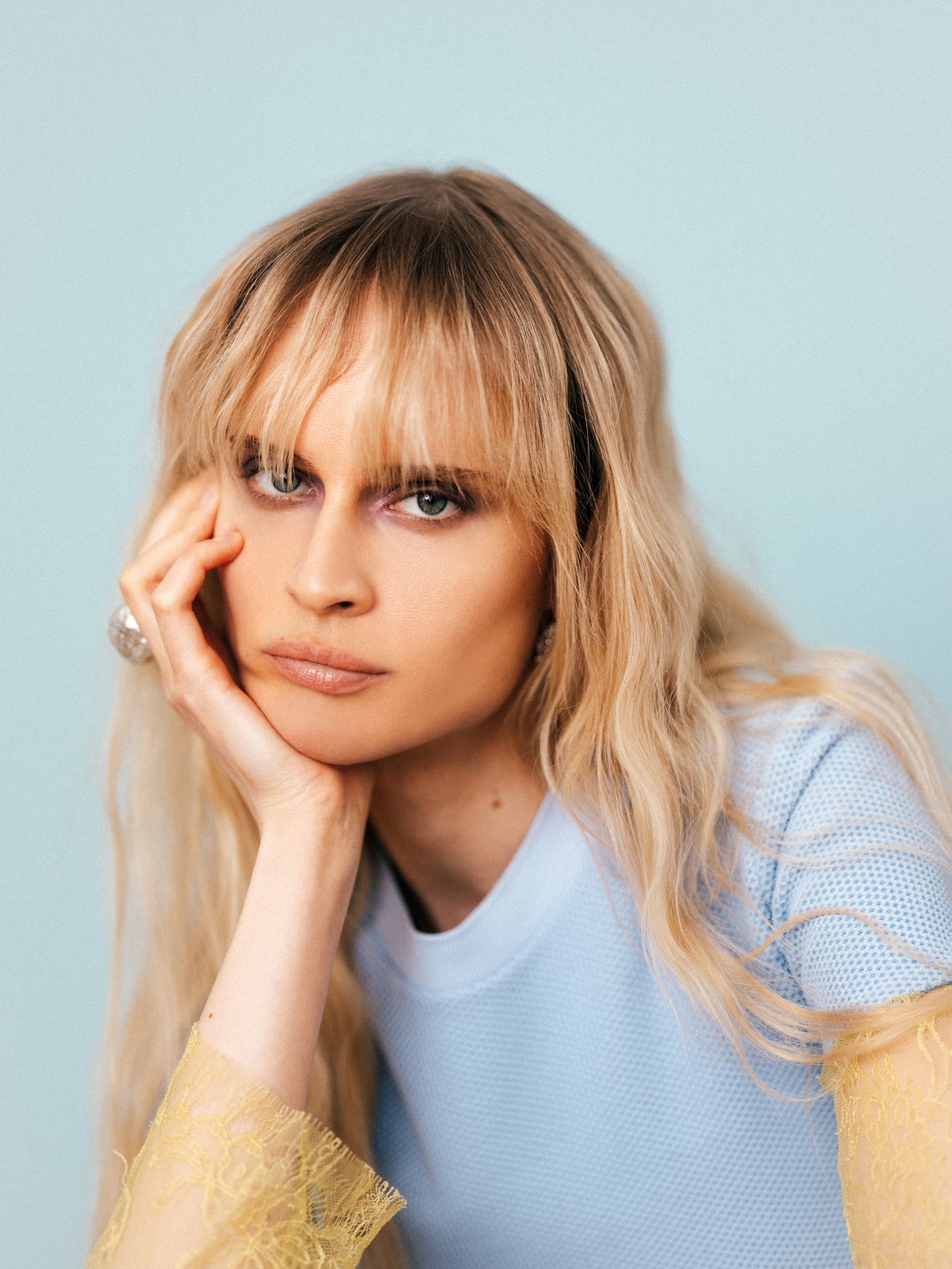 Woman with long blonde hair poses against a light blue background in a stylish outfit.