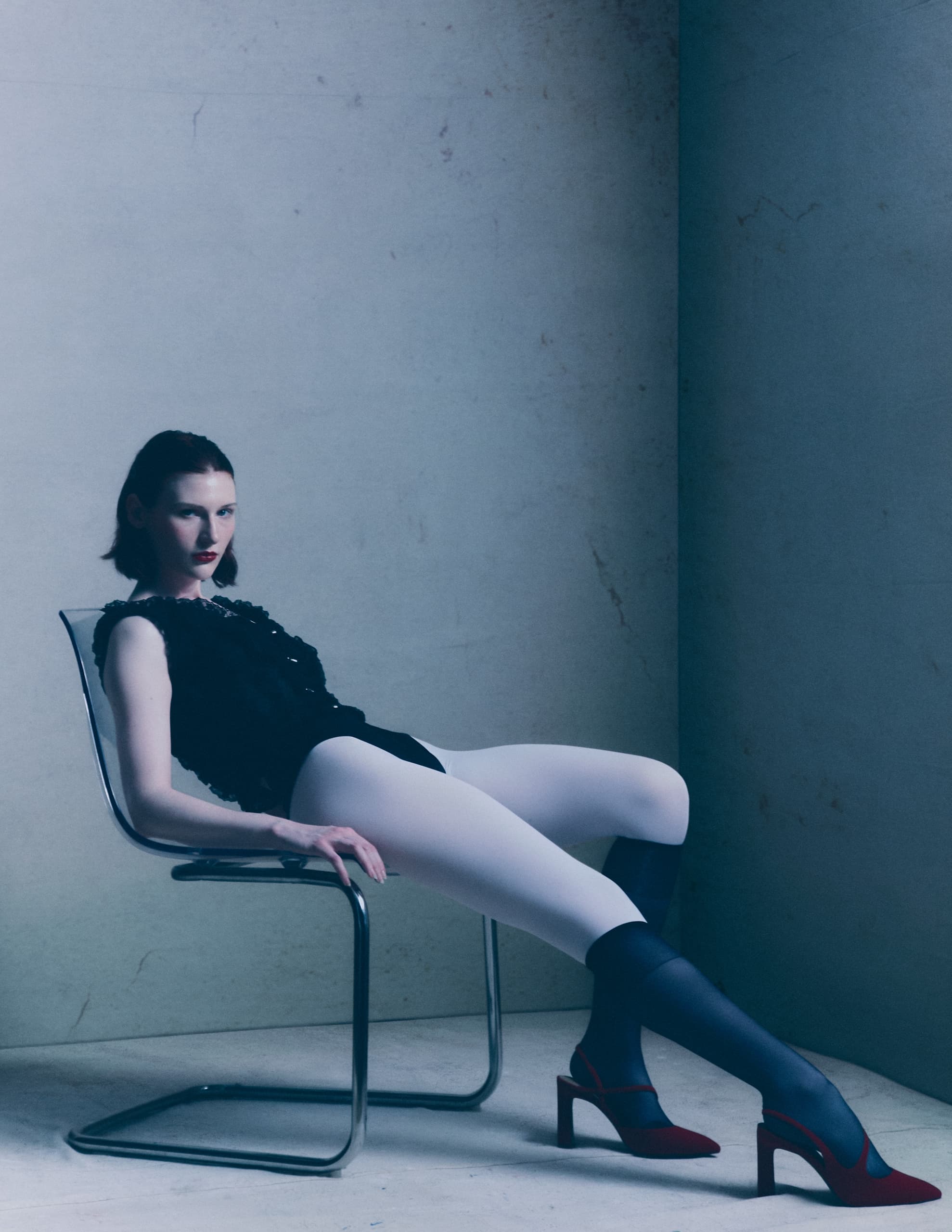 Fashion model in avant-garde outfit sitting on a modern chair in minimalist setting.