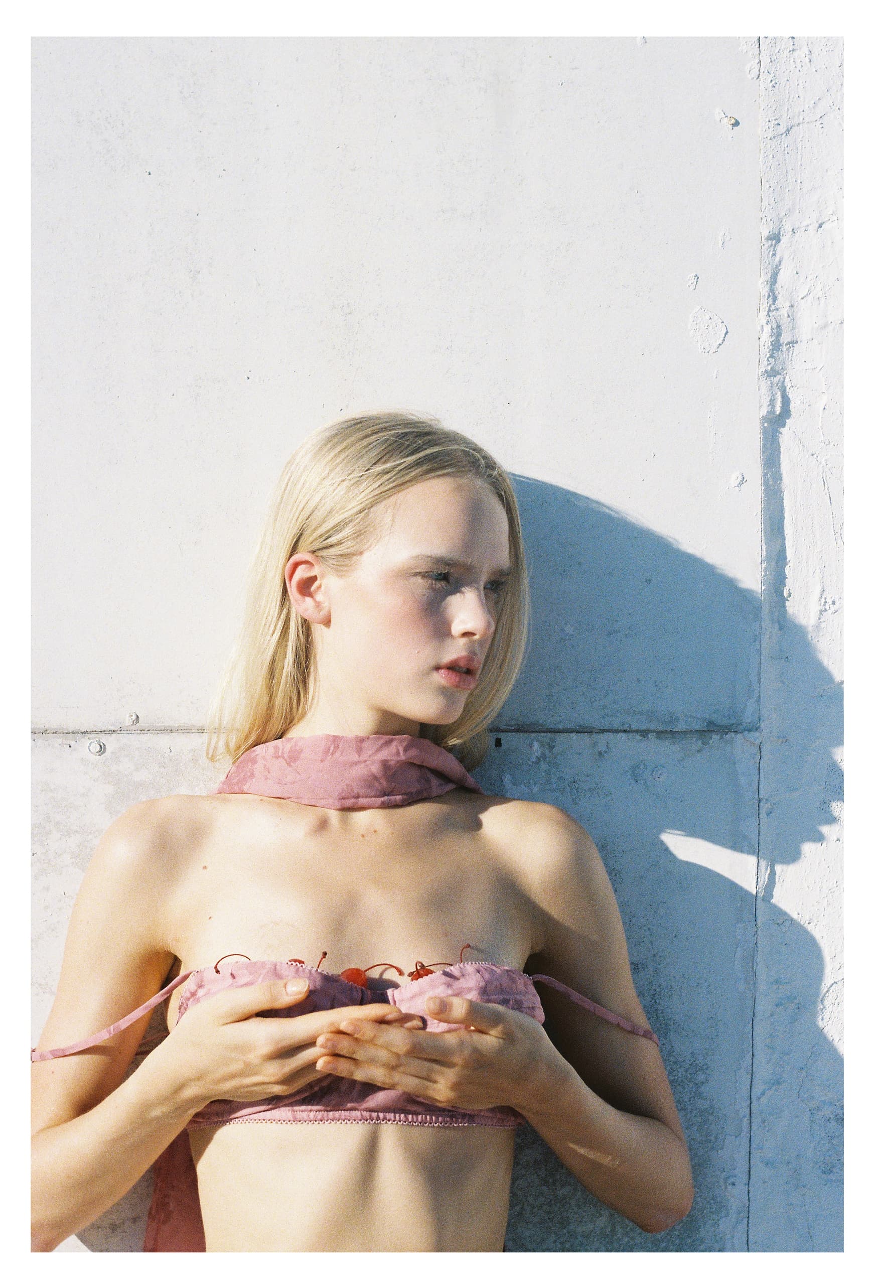 Blonde woman with pink scarf posing against a white wall in sunlight.