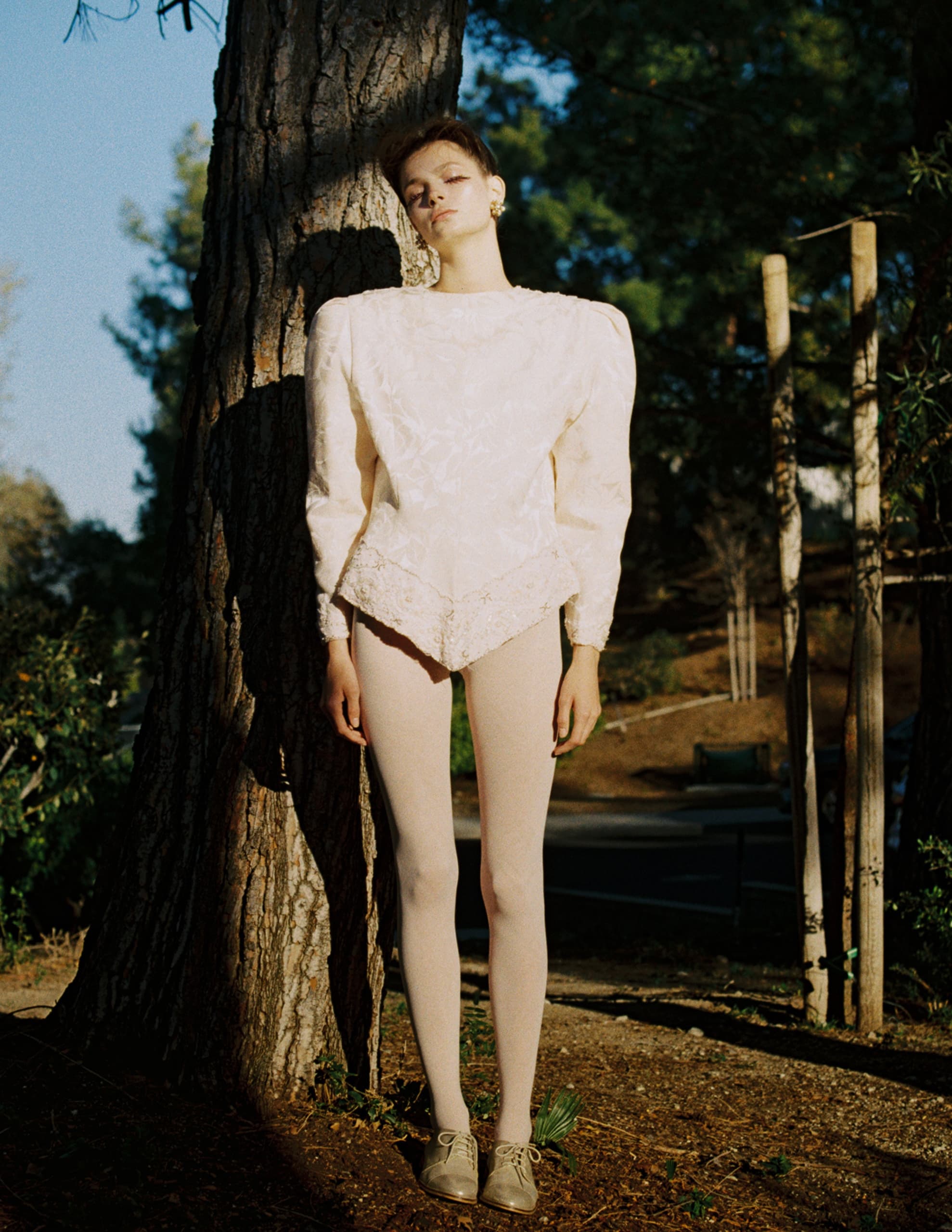 Fashion model in vintage outfit leaning against a tree in sunlight.