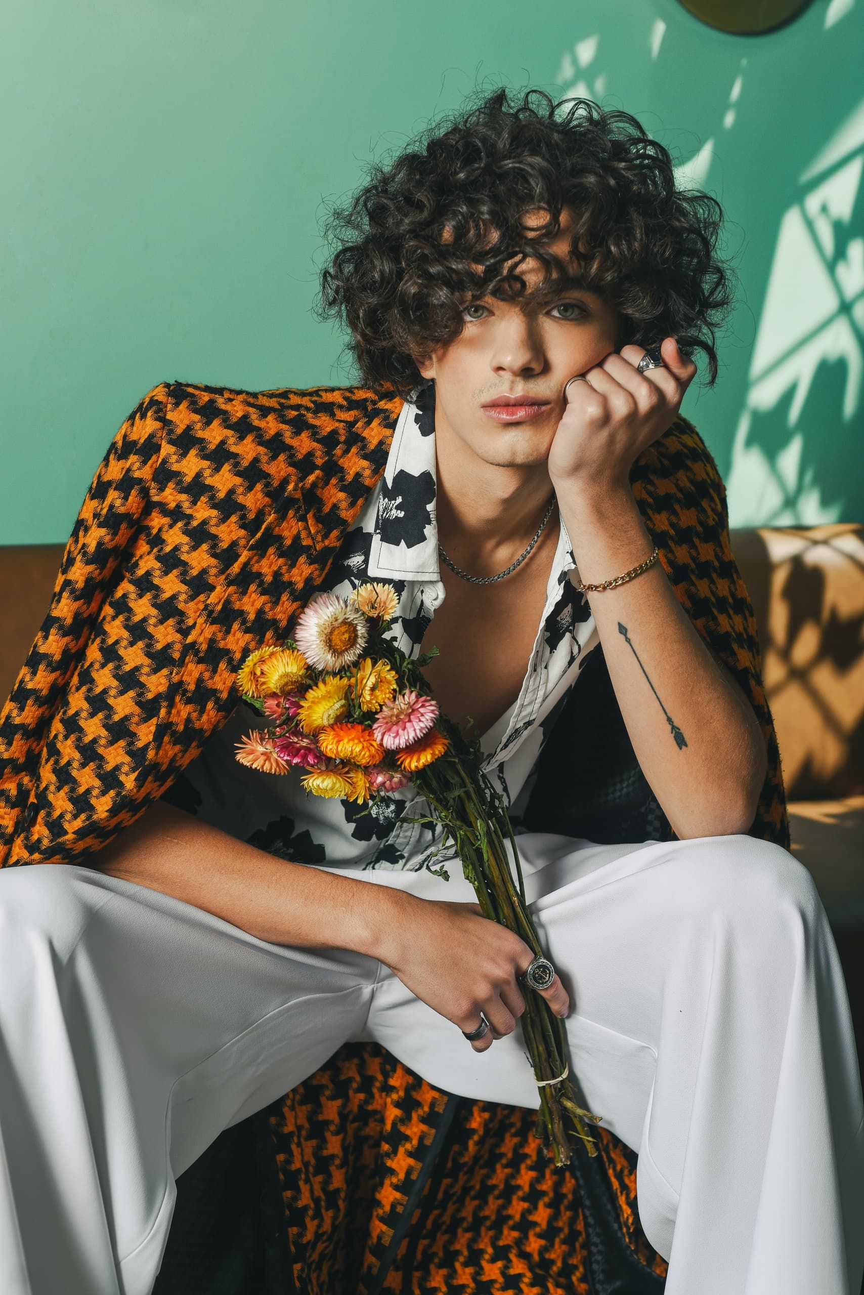 Person in stylish outfit with curly hair, holding vibrant flowers, against a teal wall.