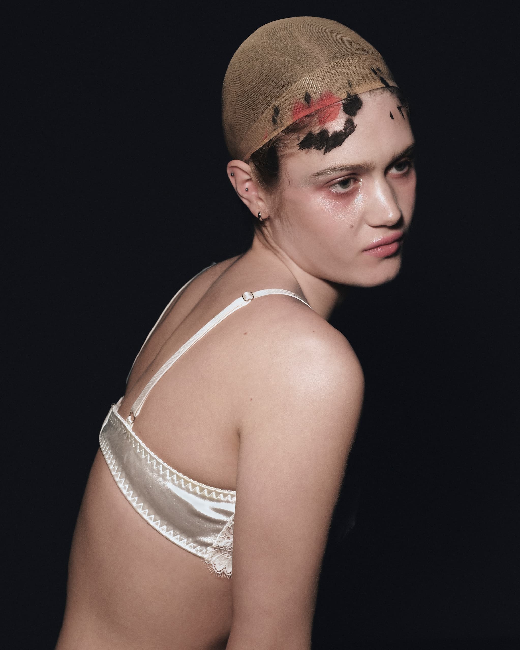 Model in lingerie with artistic face paint under sheer cap, dark background.