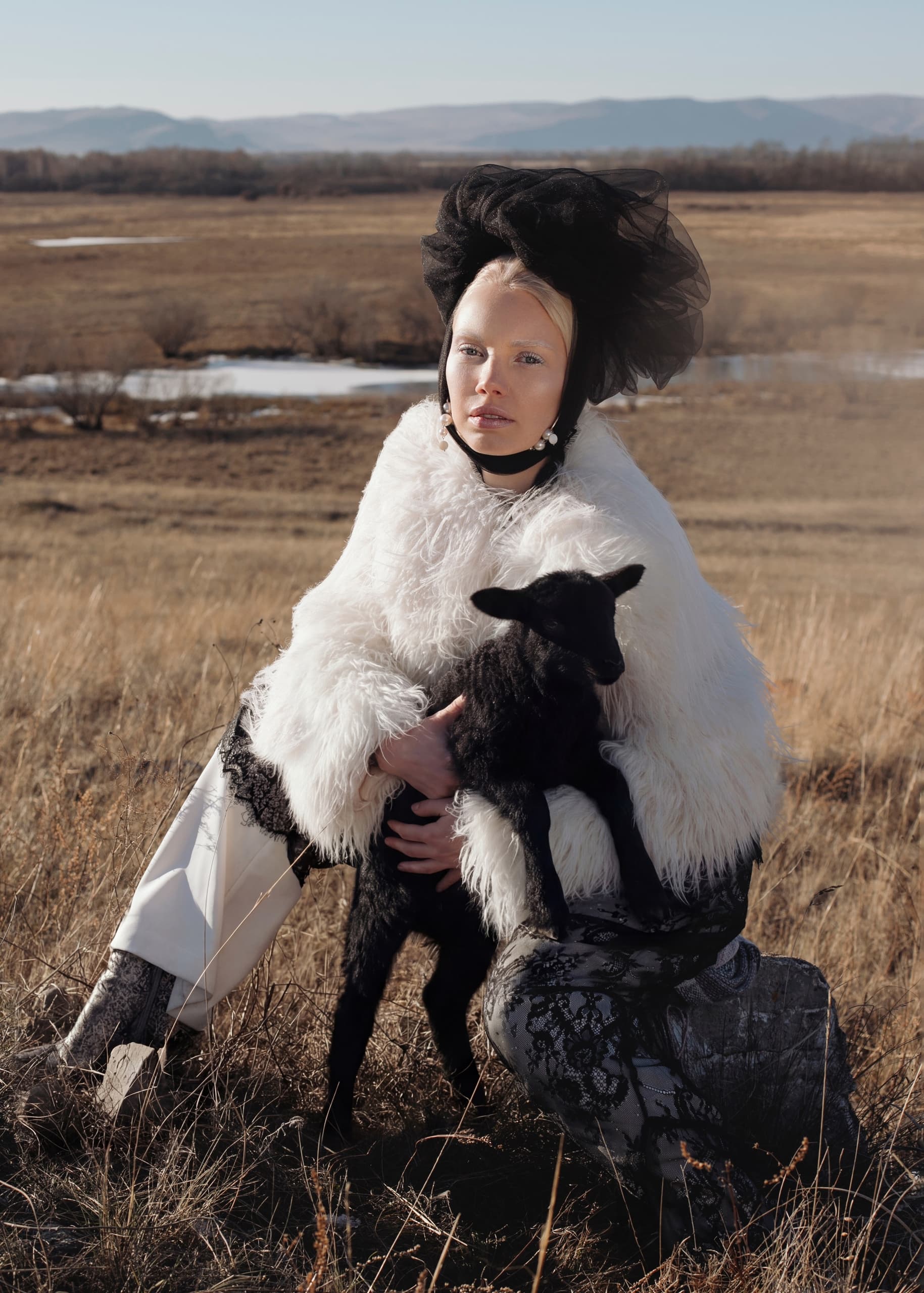 Model in white fur holding black lamb in a scenic field.