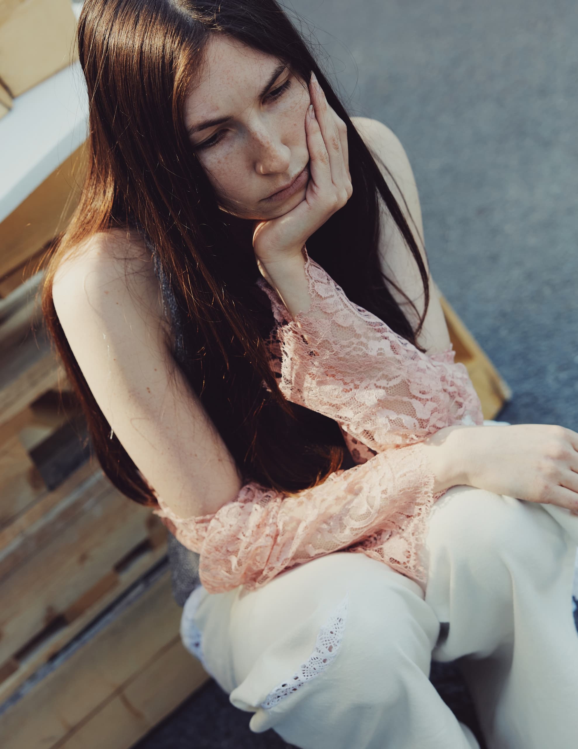 Woman in pink lace top sitting pensively outdoors.