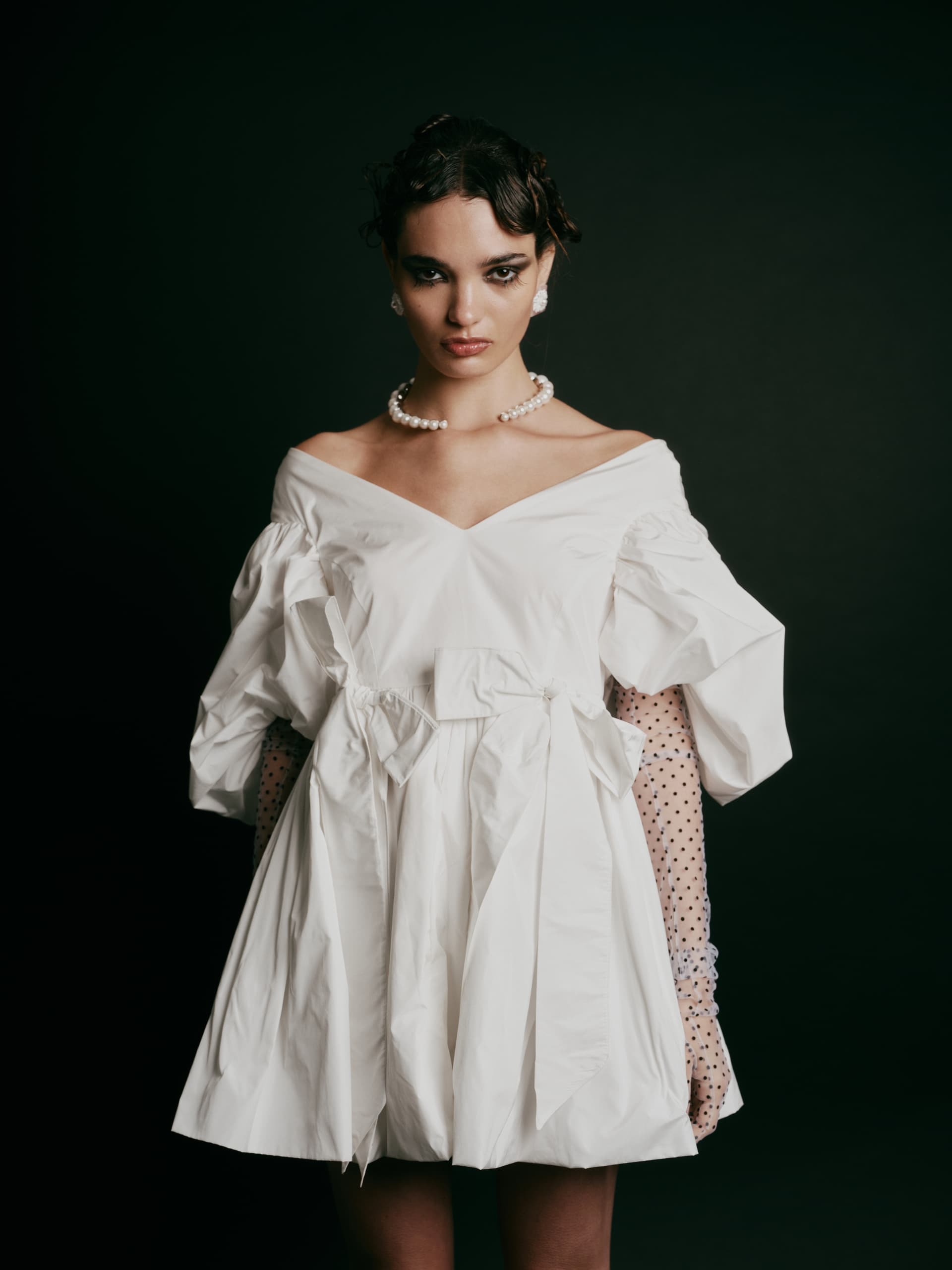 Woman in stylish white dress and pearls against dark background.