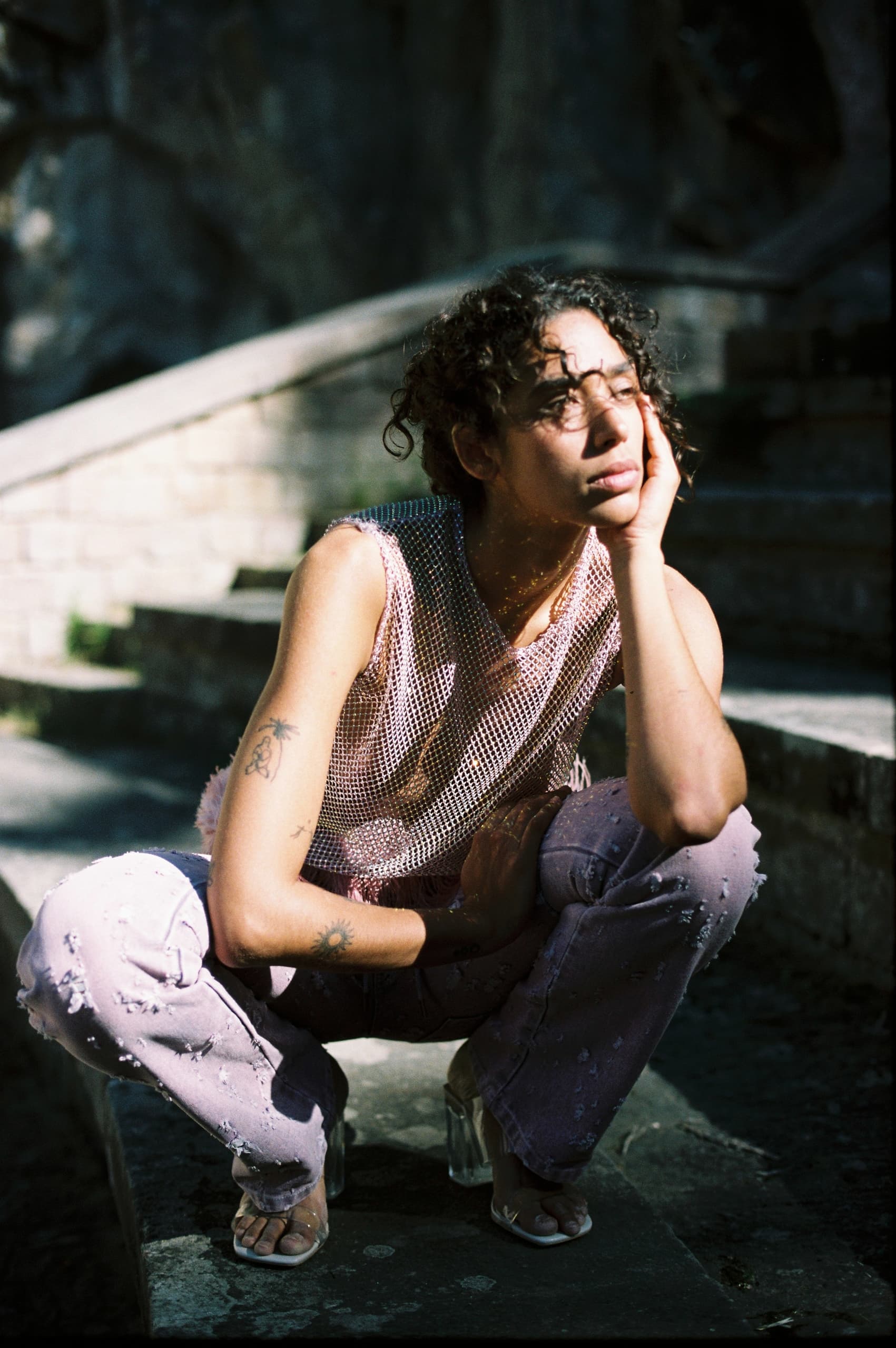 Person sitting on steps in sunlight, wearing torn jeans and a mesh top.