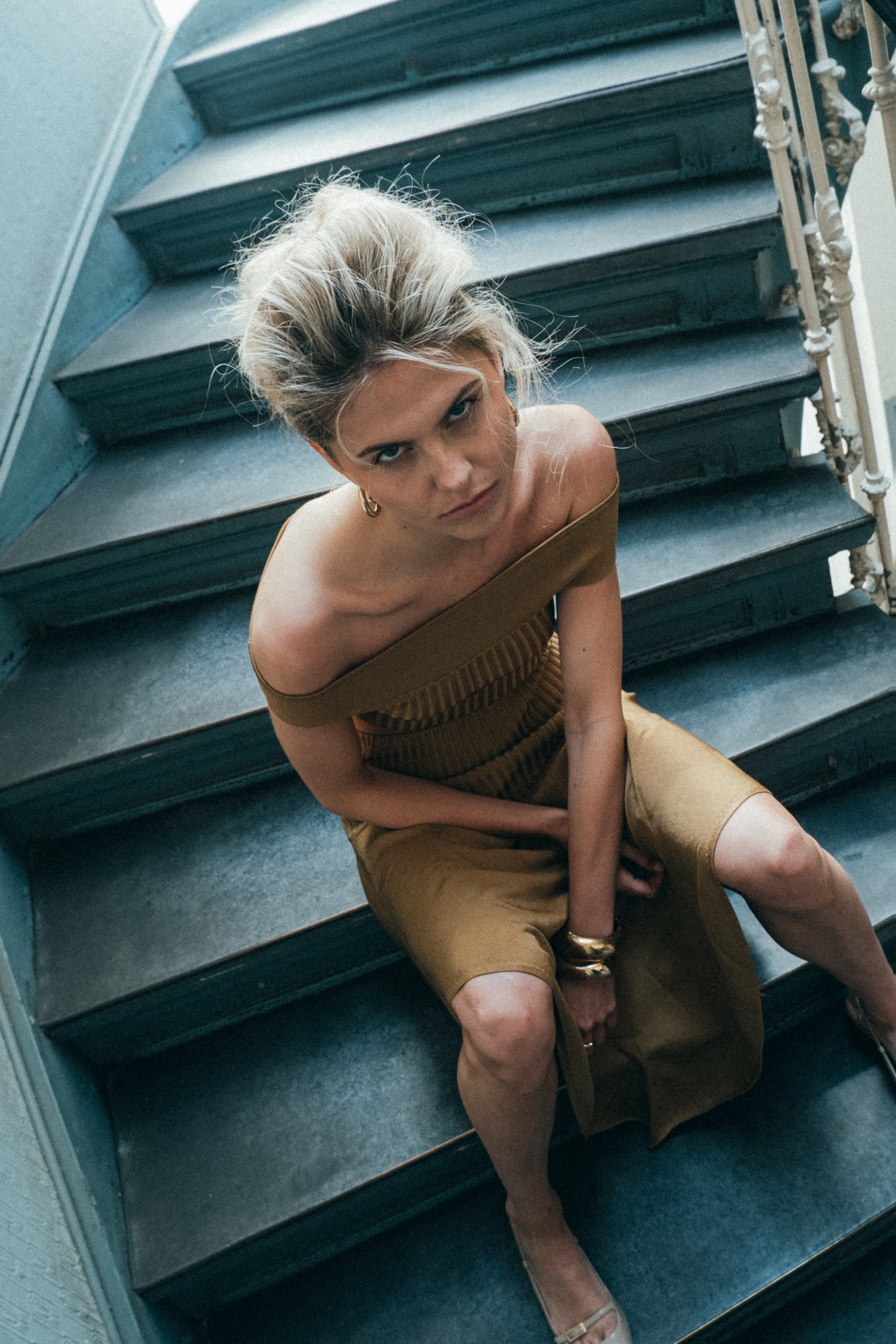 Woman in an off-shoulder dress sitting on stairs, looking up at the camera.