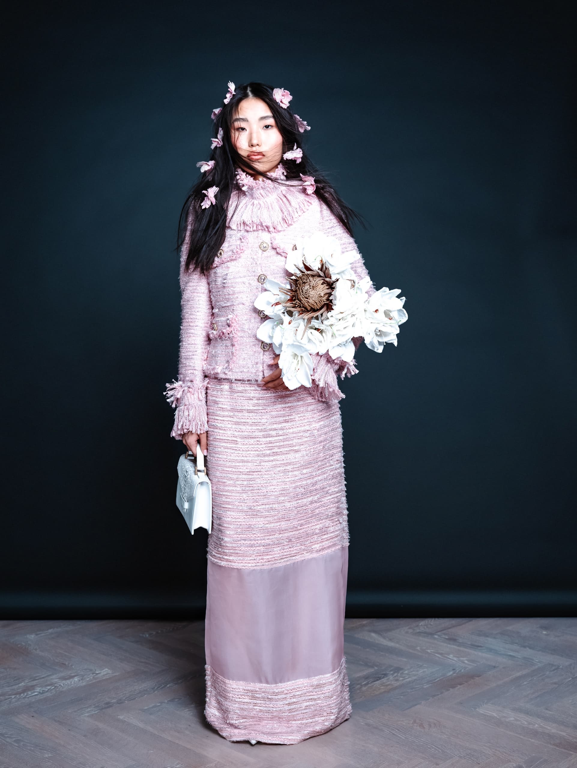 Woman in pink textured dress with flowers and white handbag against dark background.
