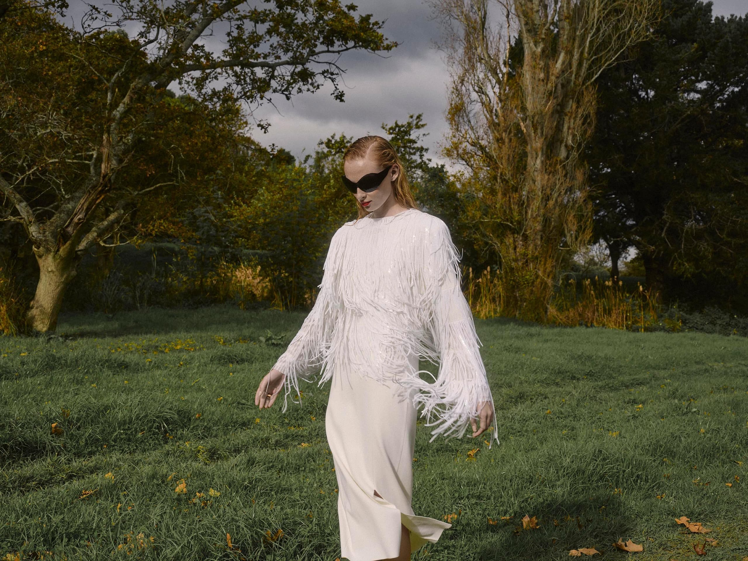 Woman in fringed white dress walking through a lush green meadow.