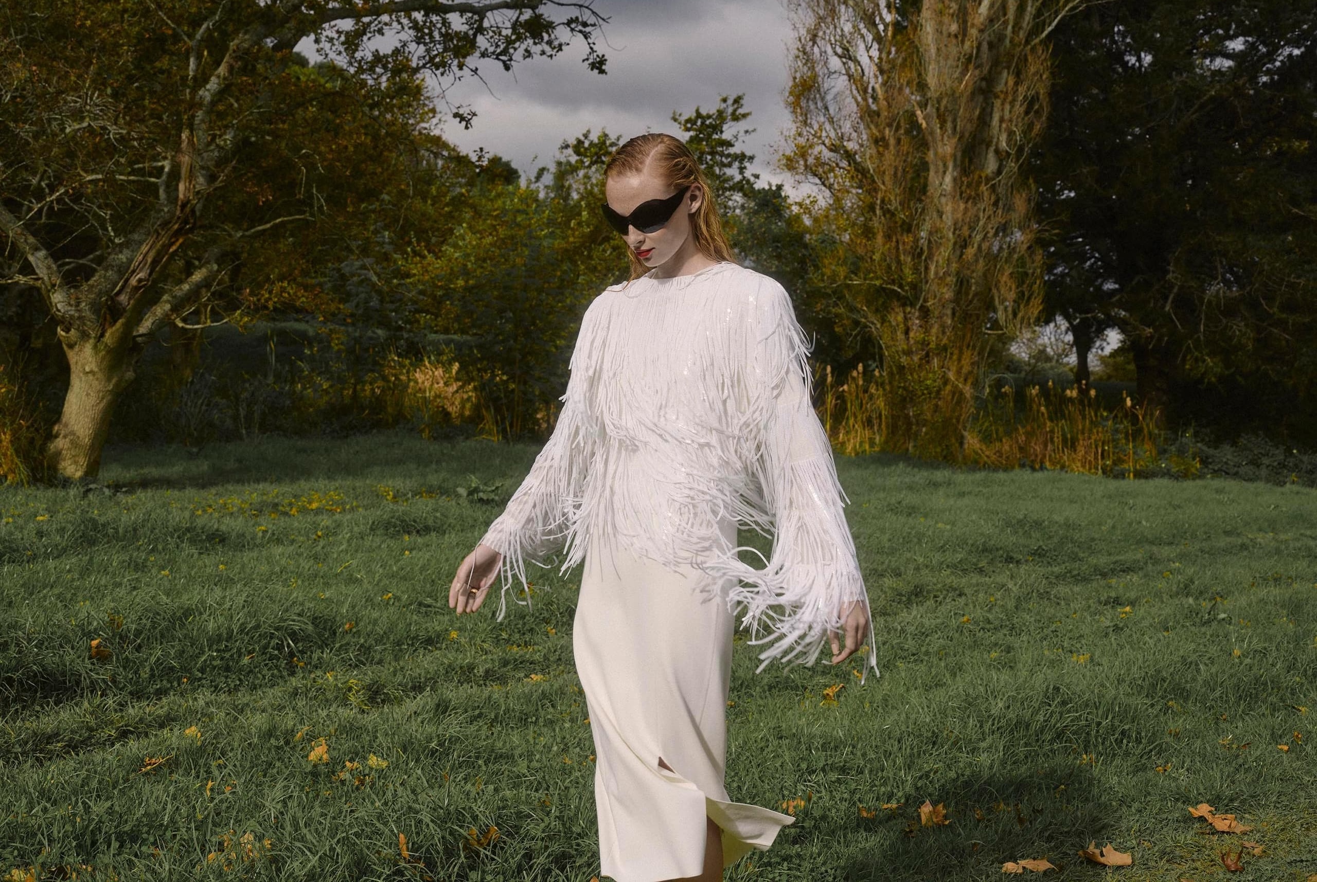 Fashionable woman in white fringe outfit walking in a green landscape.