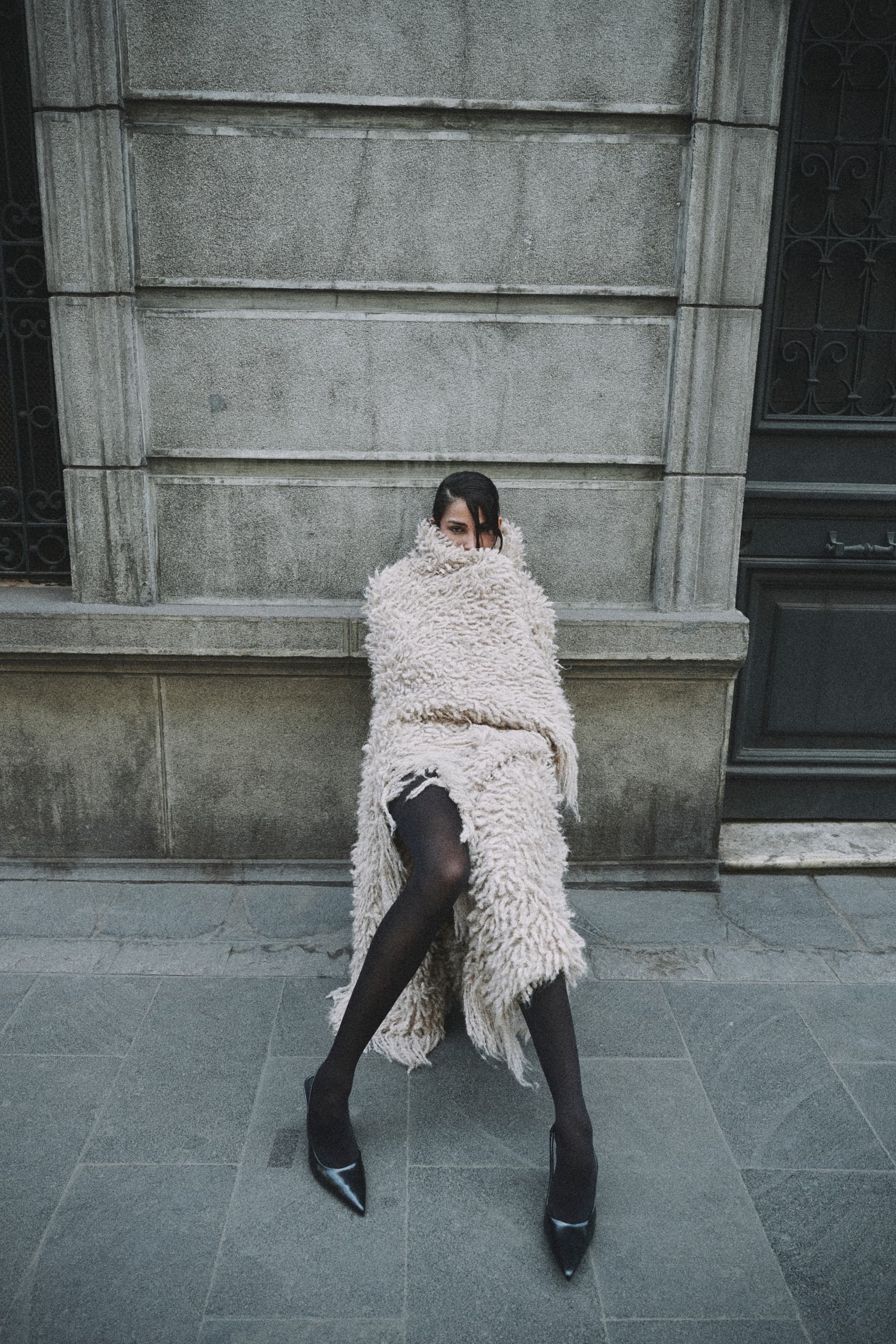 Person wrapped in a cozy, textured coat sitting against an urban wall. Fashion statement.