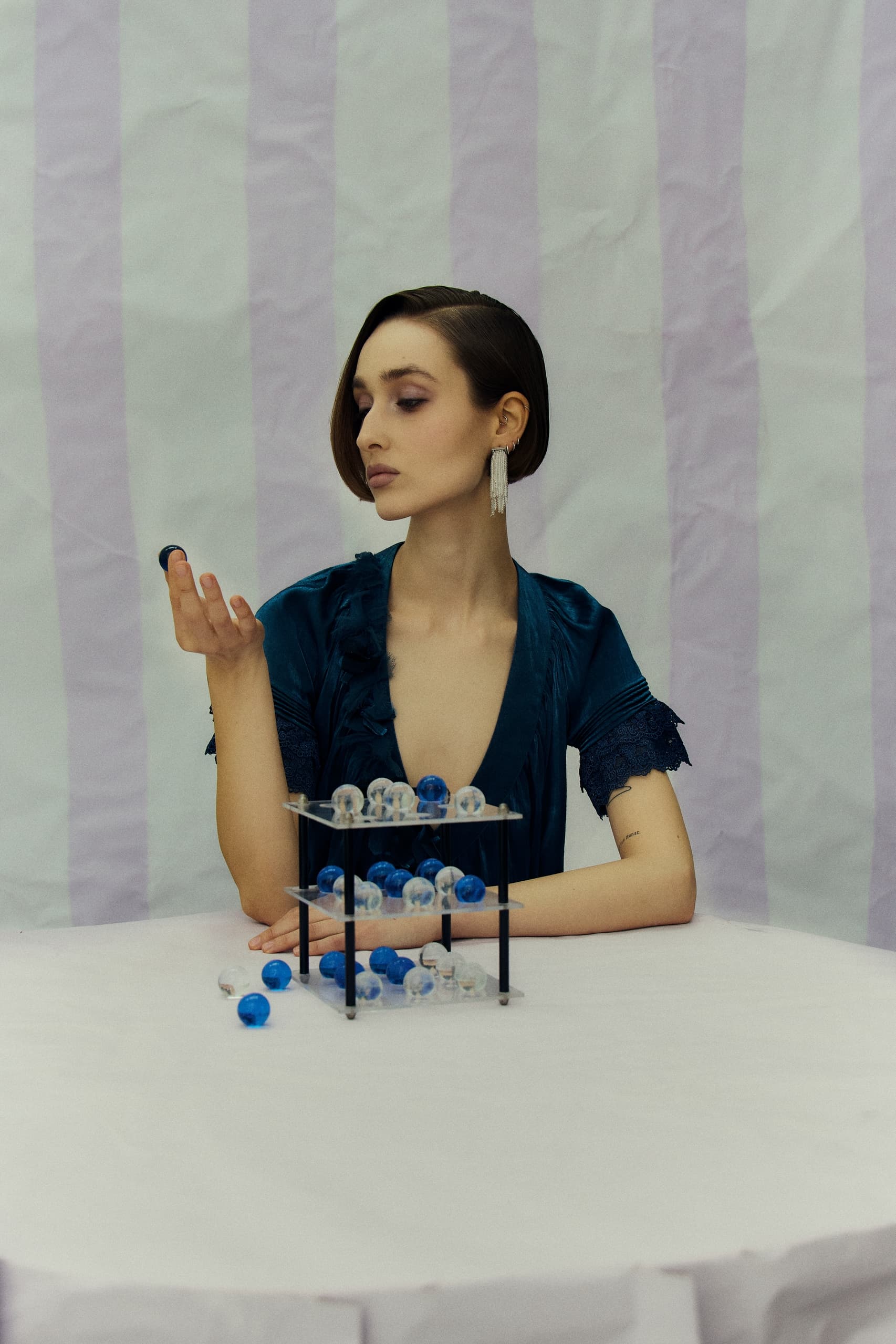 Woman in teal dress with blue spheres on table, contemplating an object.